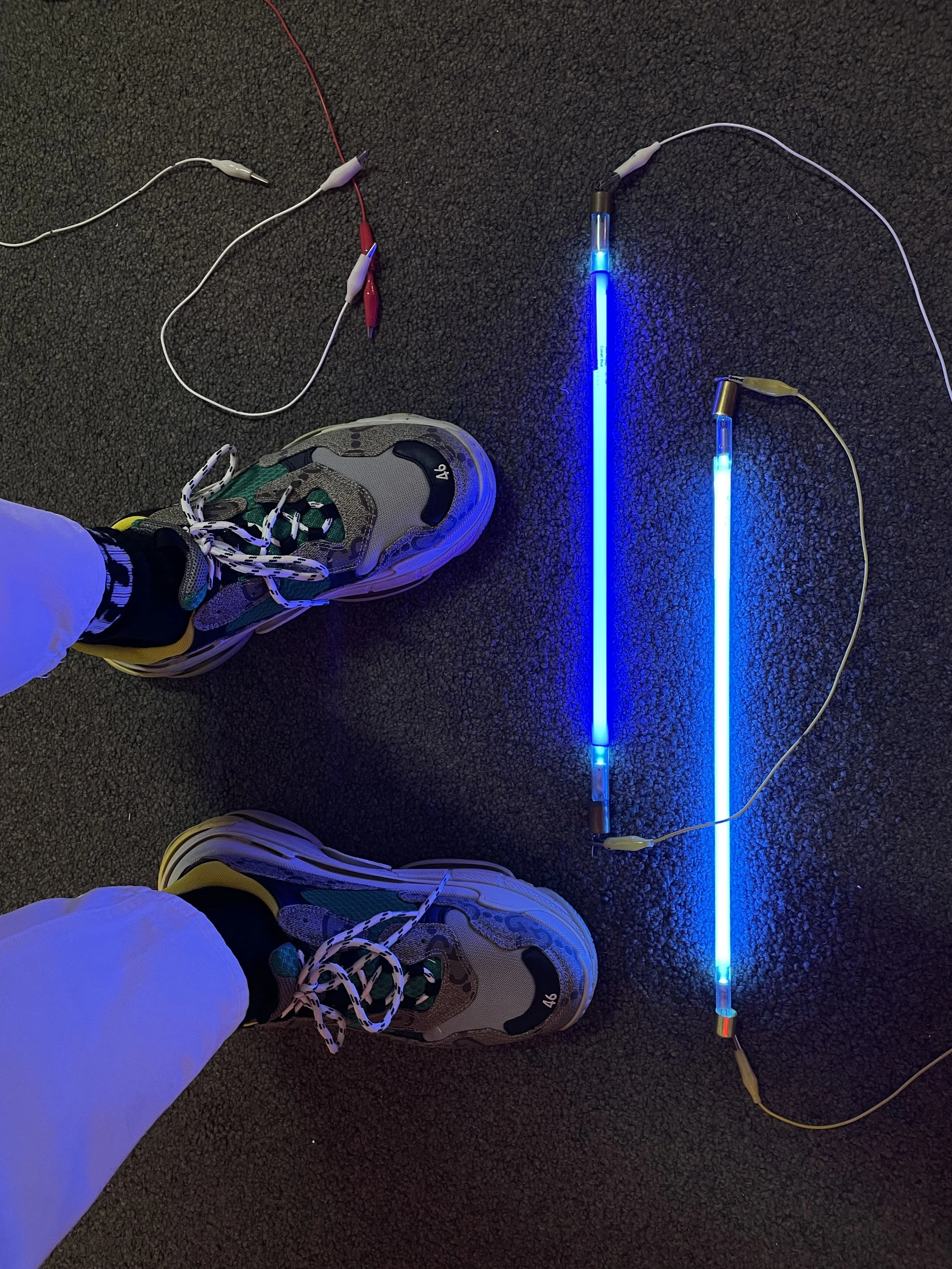 Person wearing sneakers standing on a dark carpeted floor next to two blue neon light tubes connected with red, white, and yellow wires.