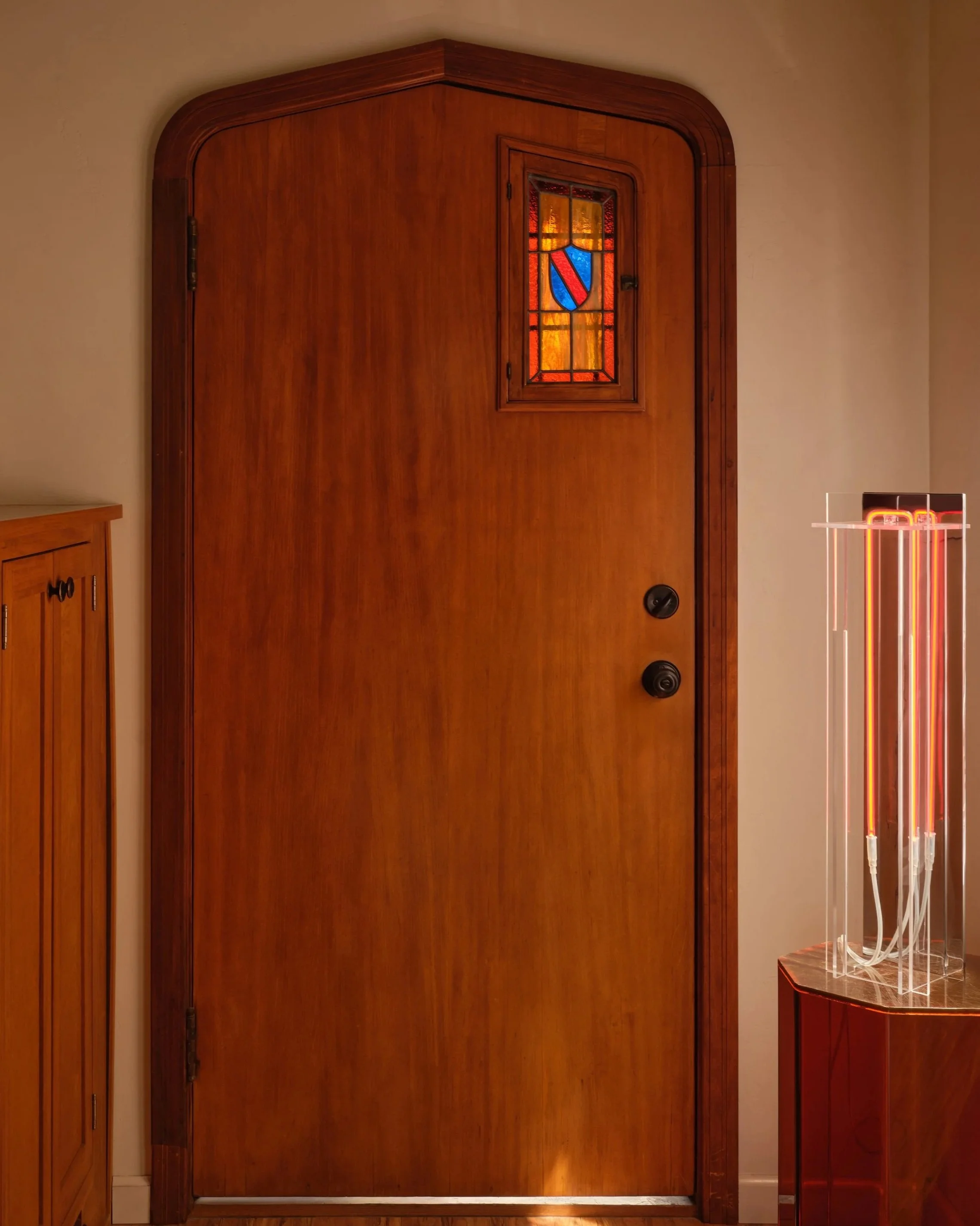 A wooden front door with a small stained glass window featuring a shield emblem, flanked by a wooden cabinet on the left and a modern neon light on the right.