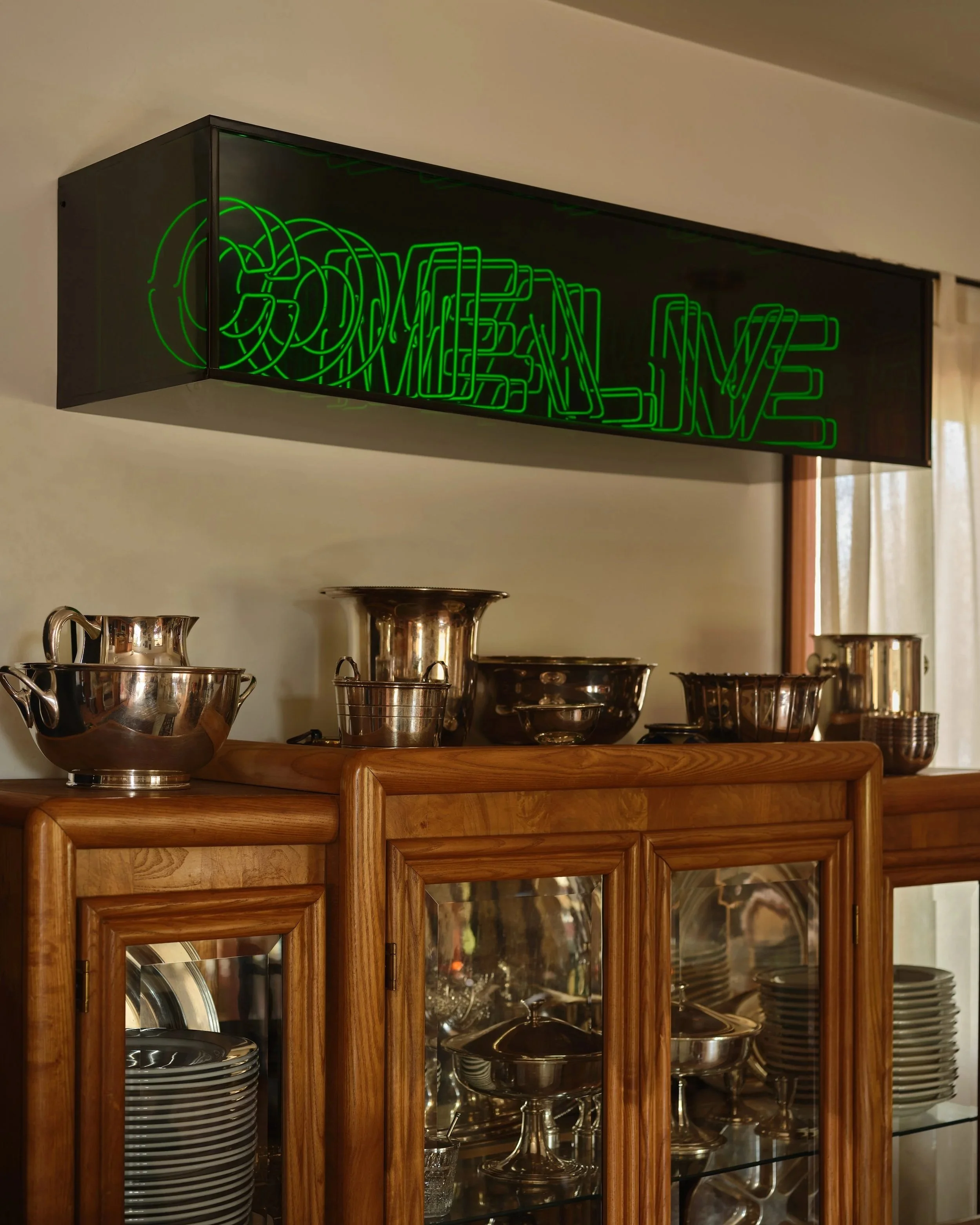 Green neon sign that says 'DRINKS ON ME' above a wooden cabinet with glass doors, displaying plates and serving bowls, with metal pots and bowls on top of the cabinet.