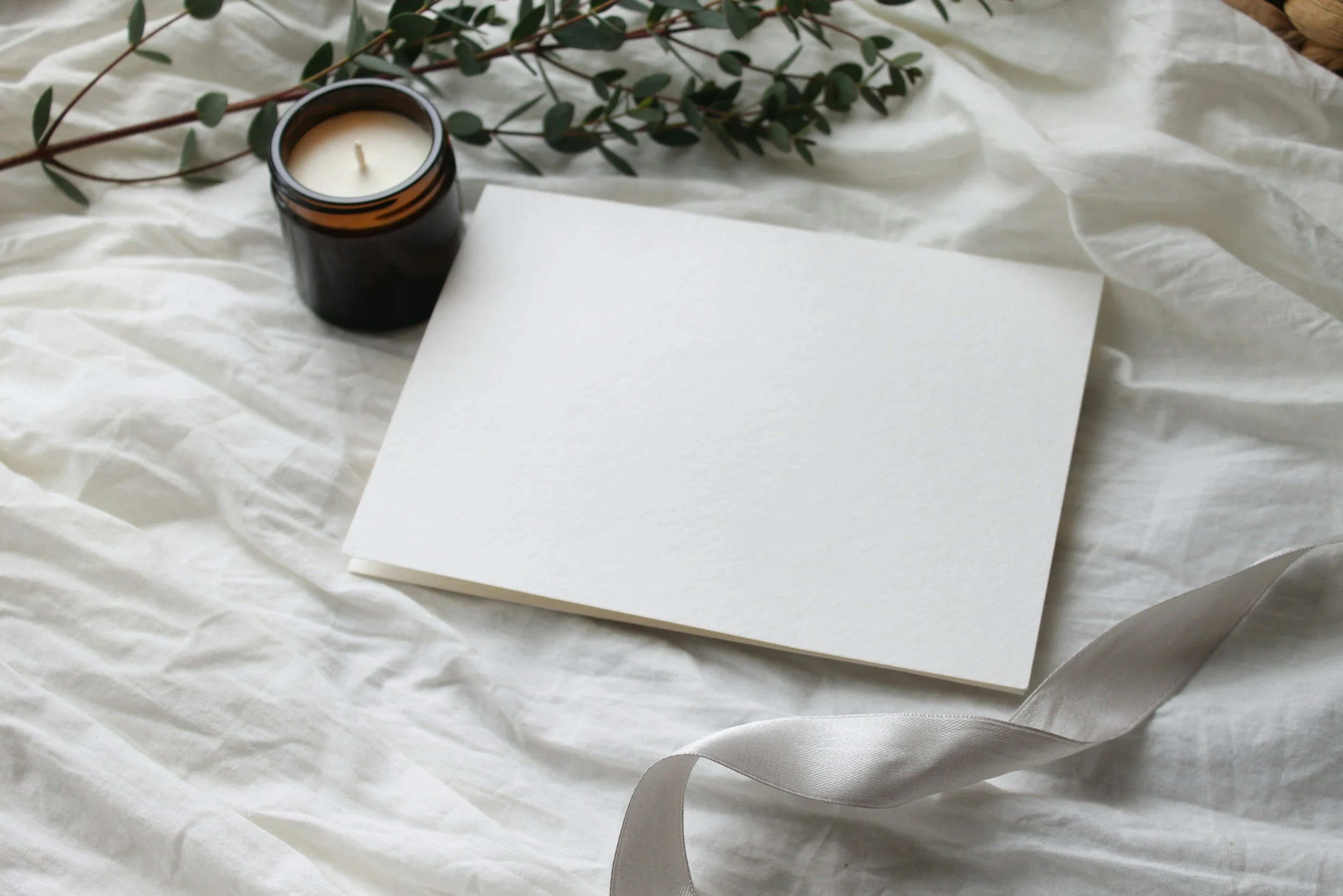 A white candle in a dark glass jar, a white sheet of paper, and a sprig of greenery on crumpled white fabric.