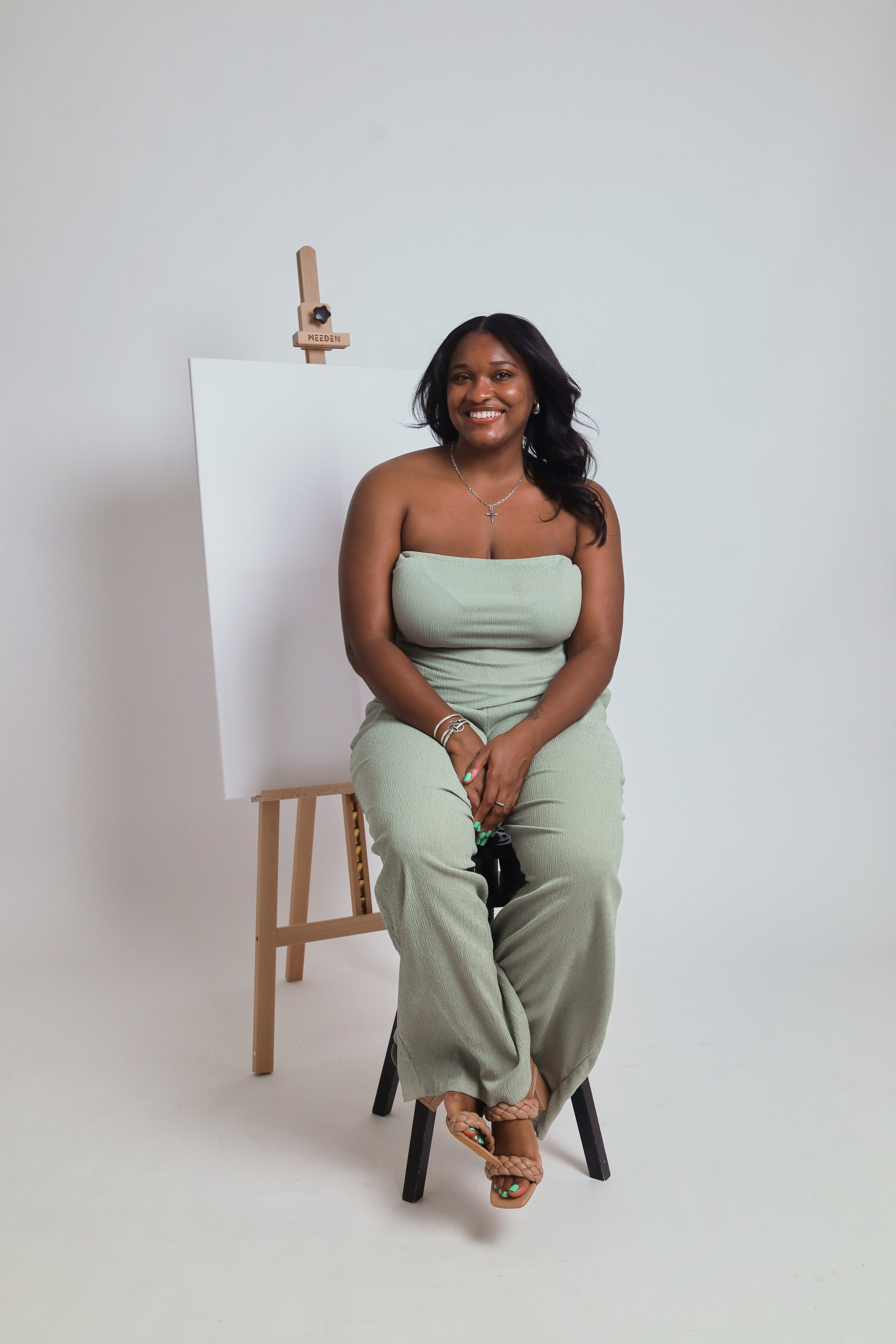 A woman with dark, wavy hair smiling while sitting on a stool in front of a white background, wearing a light green strapless jumpsuit, tan sandals, and jewelry, with an easel and canvas behind her.