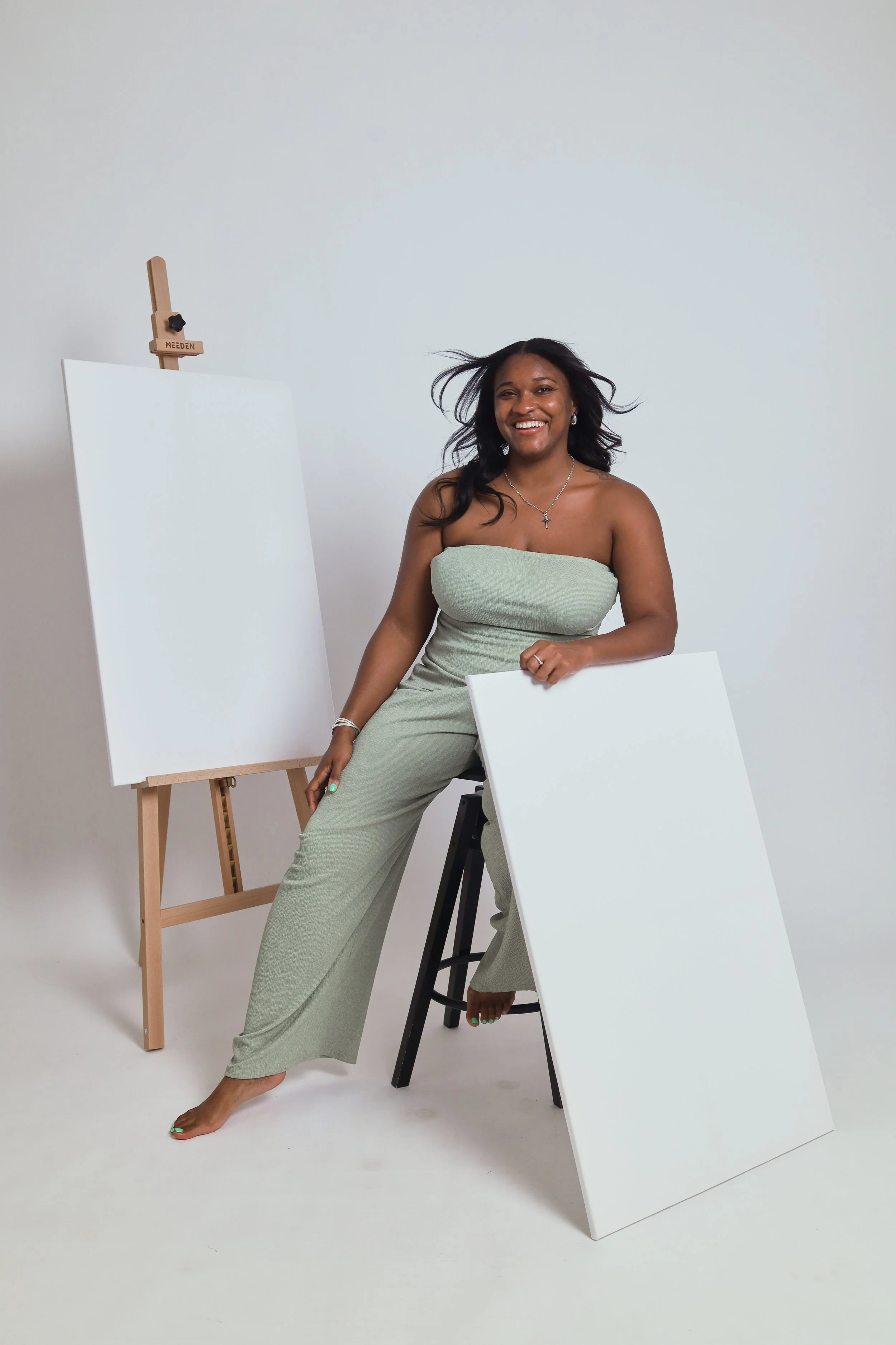 A woman with long dark hair, smiling, wearing a strapless light green outfit, sitting on a black stool with one leg crossed over the other, in a studio with white walls. There are blank canvases and an easel behind her.