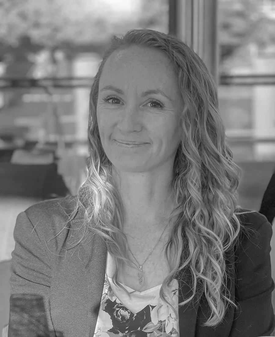 A woman with long curly hair, smiling, wearing a blazer over a floral top with a necklace, sitting indoors with a blurred background.