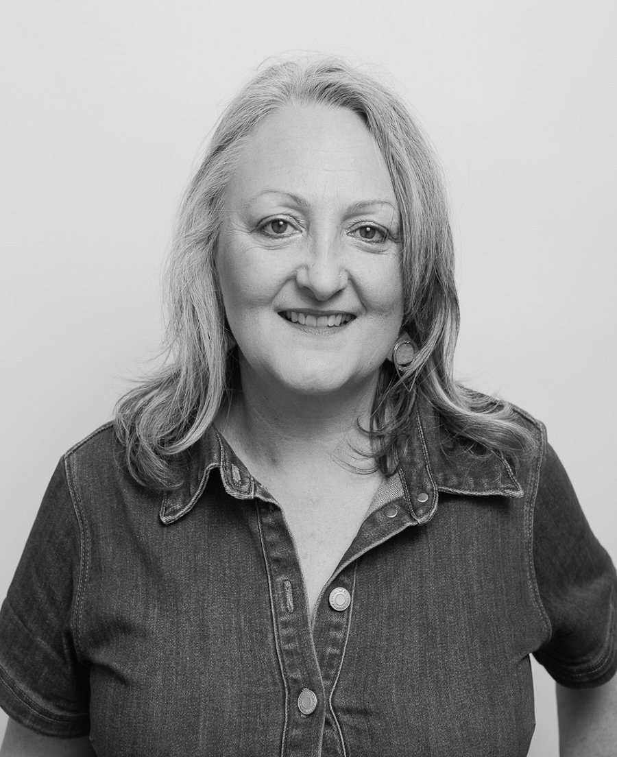 A black and white portrait of a smiling woman with shoulder-length wavy hair, wearing a denim shirt, against a plain light background.