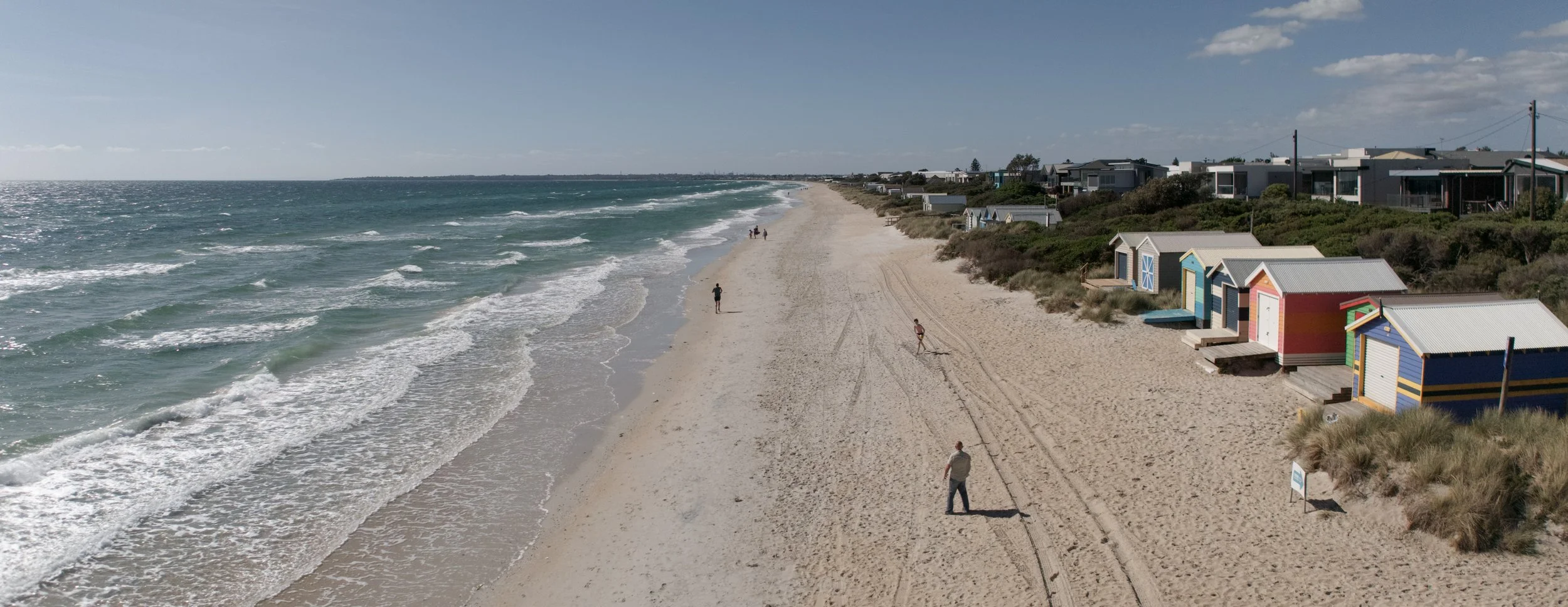 A wide aerial view of Chelsea Beach in Victoria, showing waves rolling onto the sand, colourful beach boxes along the shoreline, and a few people walking near the water.