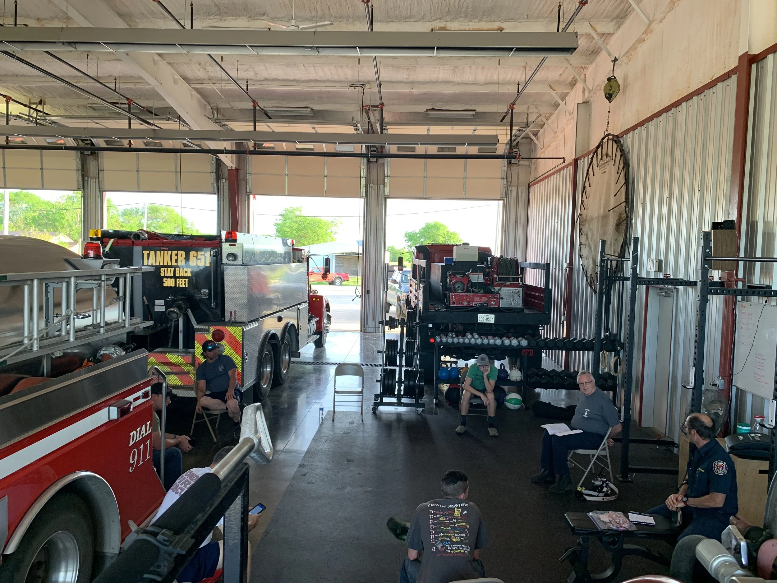 Inside a fire station garage with fire trucks, equipment, and people sitting and talking, some with notebooks and phones.