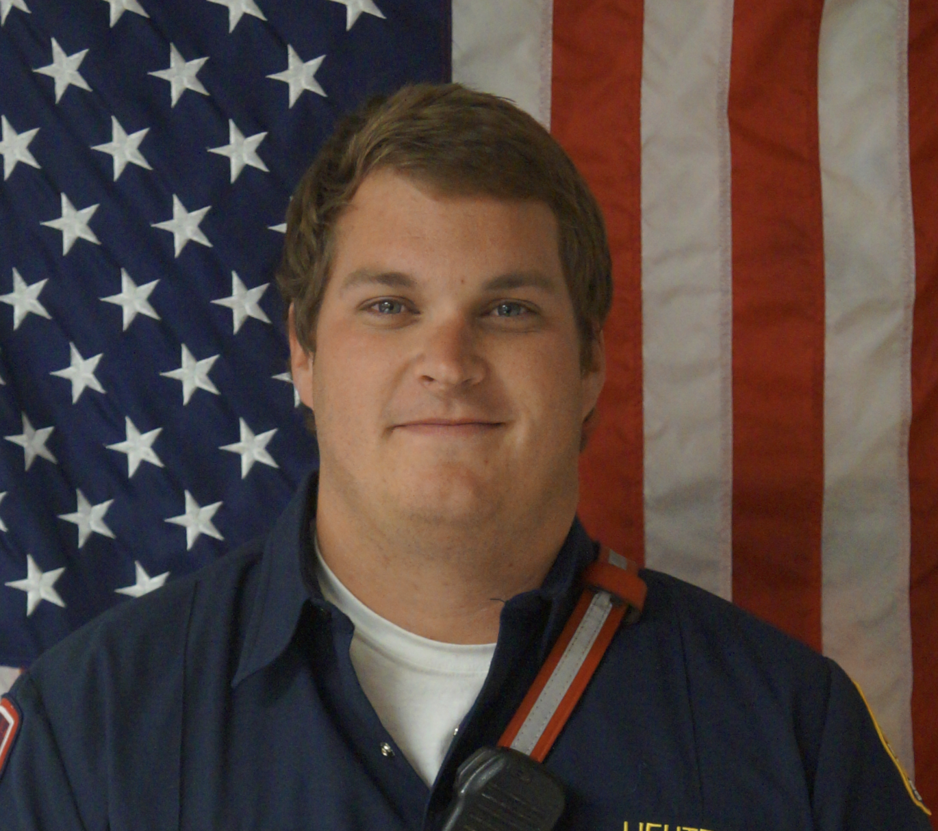 A young man in a fire department uniform standing in front of a large American flag.