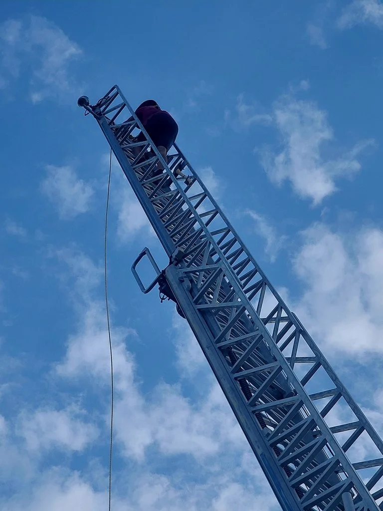 Person on a tall fire truck ladder against blue sky with clouds.