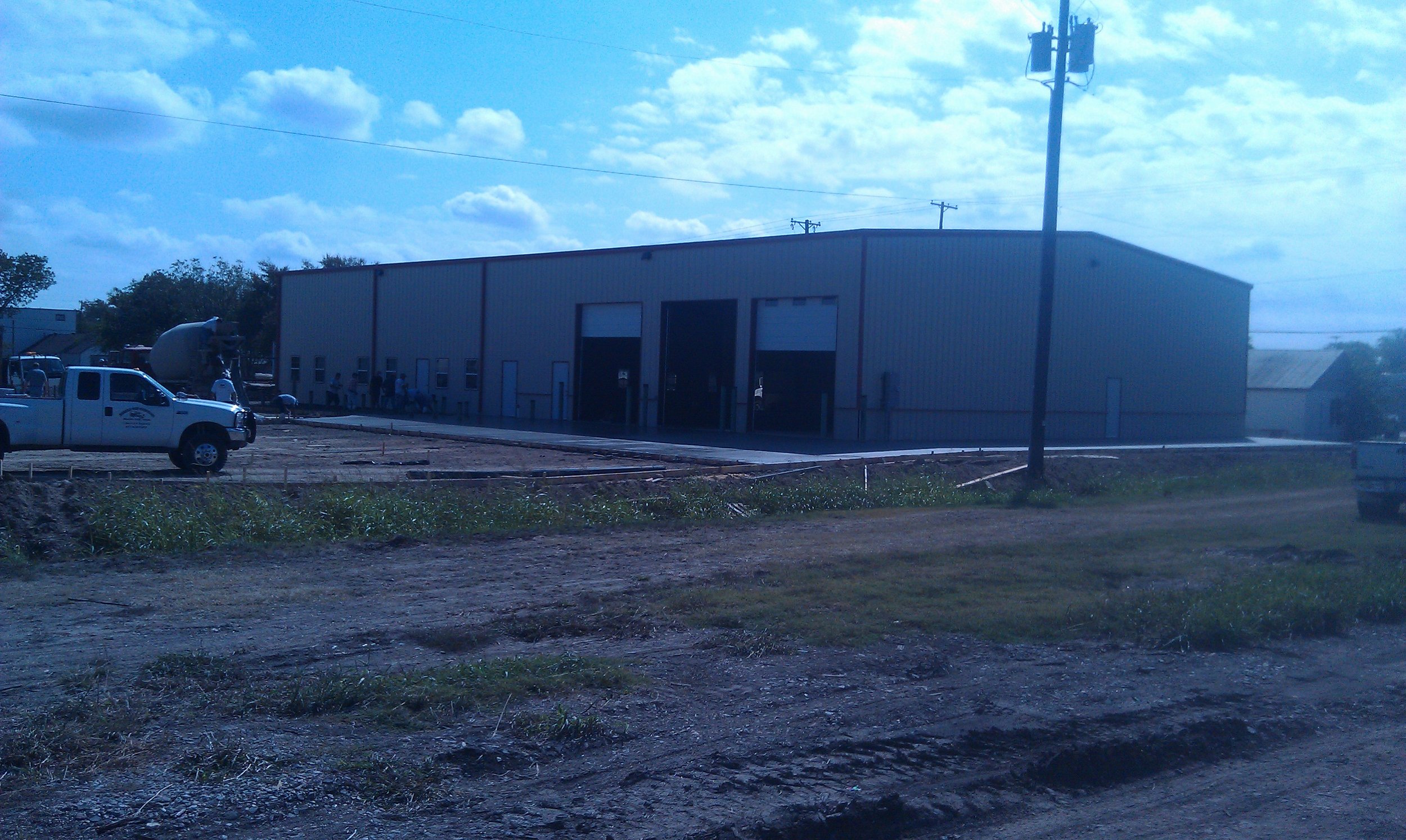 Construction site with a large metal building, a white truck, and a cement mixer, with a dirt road and green grass in the foreground and a cloudy blue sky above.