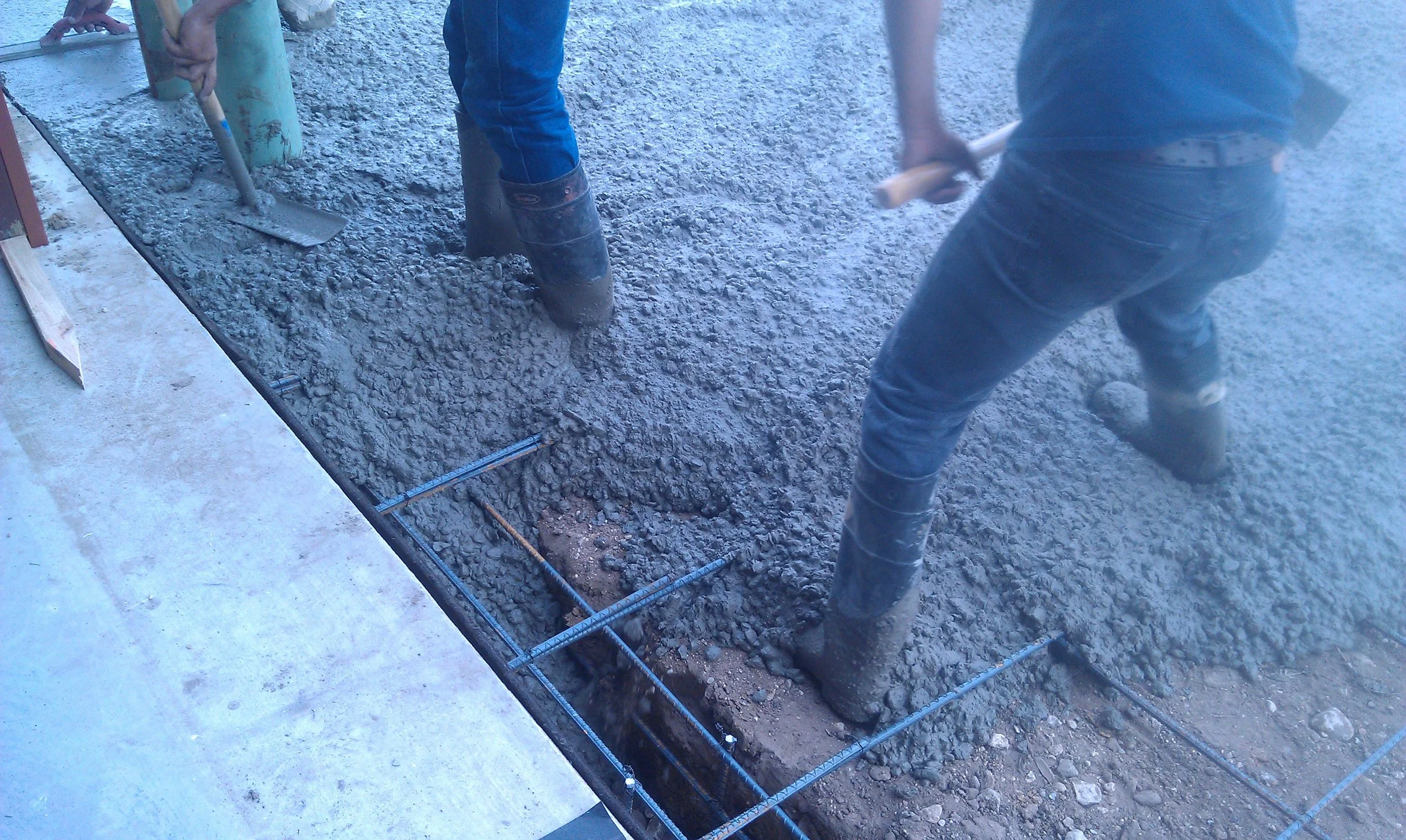 Workers are pouring and leveling concrete in a construction site, with visible rebar framework and gravel footing.