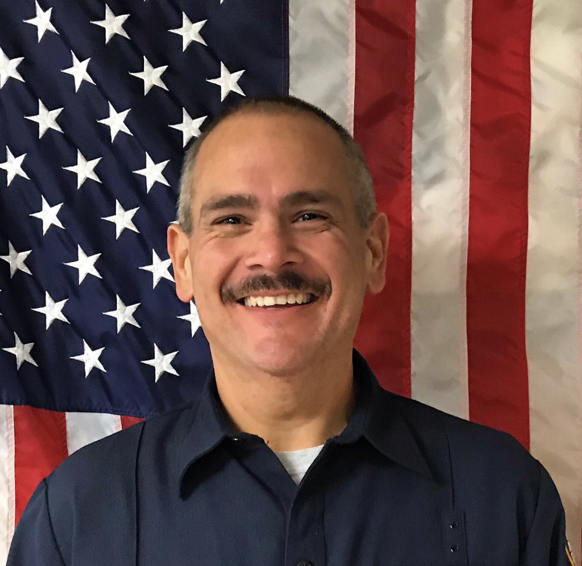 A smiling man dressed in a dark blue uniform standing in front of an American flag.