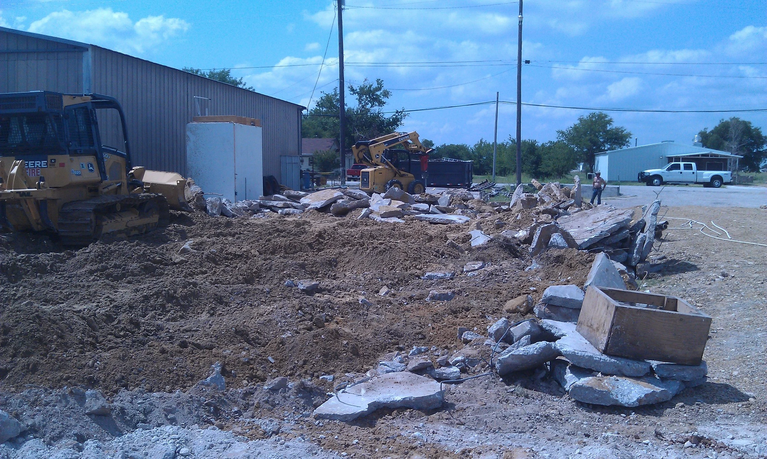 Construction site with bulldozer and backhoe moving debris and rocks, empty wooden planter, industrial buildings, utility poles, and trucks in the background under a partly cloudy sky.