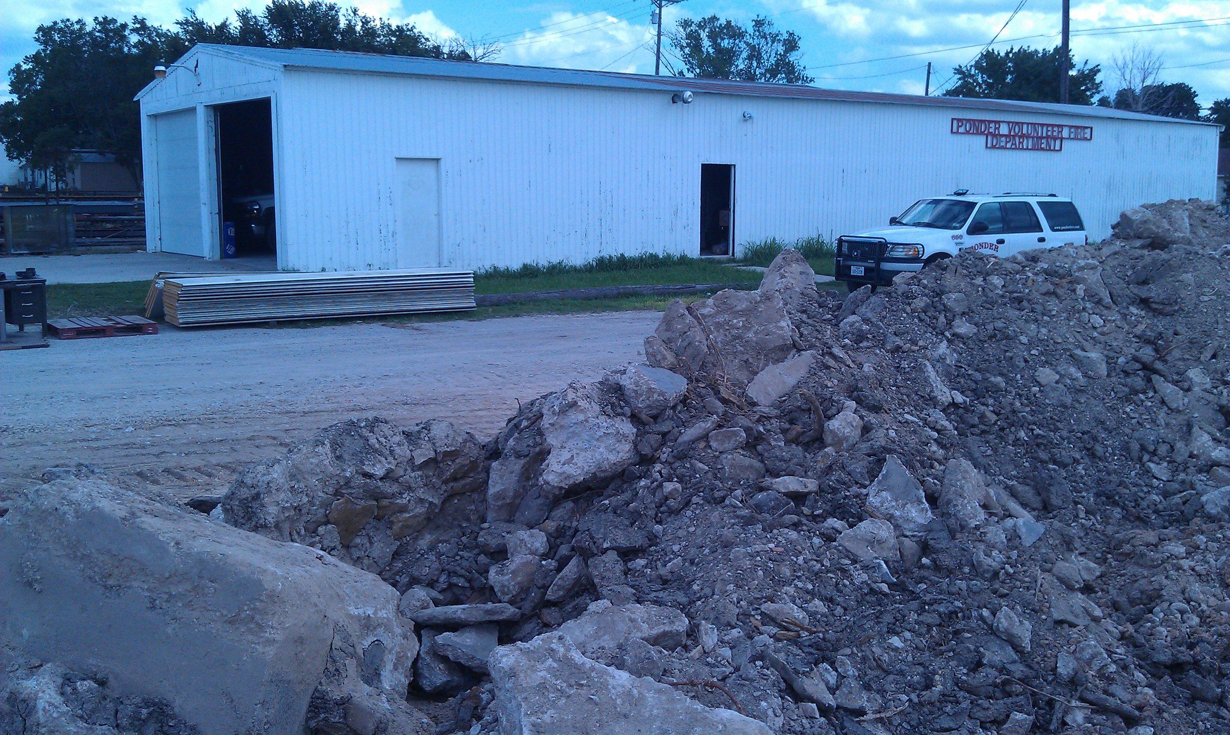 Construction site with a large pile of dirt and rocks in the foreground, a white storage building labeled 'Ponder Volunteer Fire Department' in the background, a police vehicle parked next to the building, and construction materials and tools nearby.