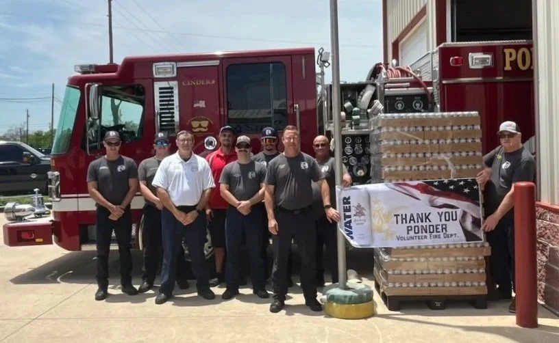 Group of firefighters and volunteers standing in front of a fire truck, holding a banner that reads 'Thank You Ponder Volunteer Fire Dept,' surrounded by supplies.