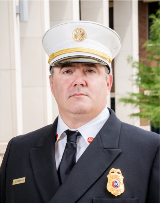 A formal portrait of a firefighter in uniform, standing outdoors with buildings and greenery in the background.