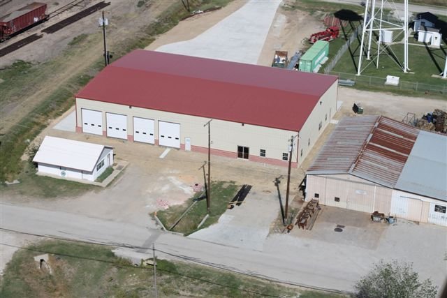Aerial view of a large beige building with a red roof, four garage doors, a small white shed nearby, and an older metal-roofed structure to the side, surrounded by utility poles and a dirt road.