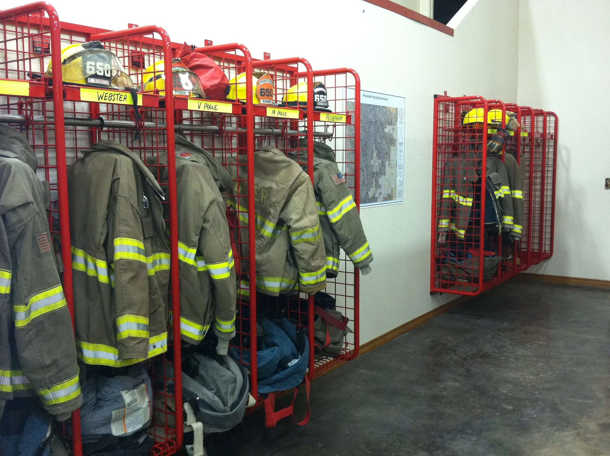 Firefighter gear, including jackets, helmets, and bags, organized on red metal racks in a fire station storage room.