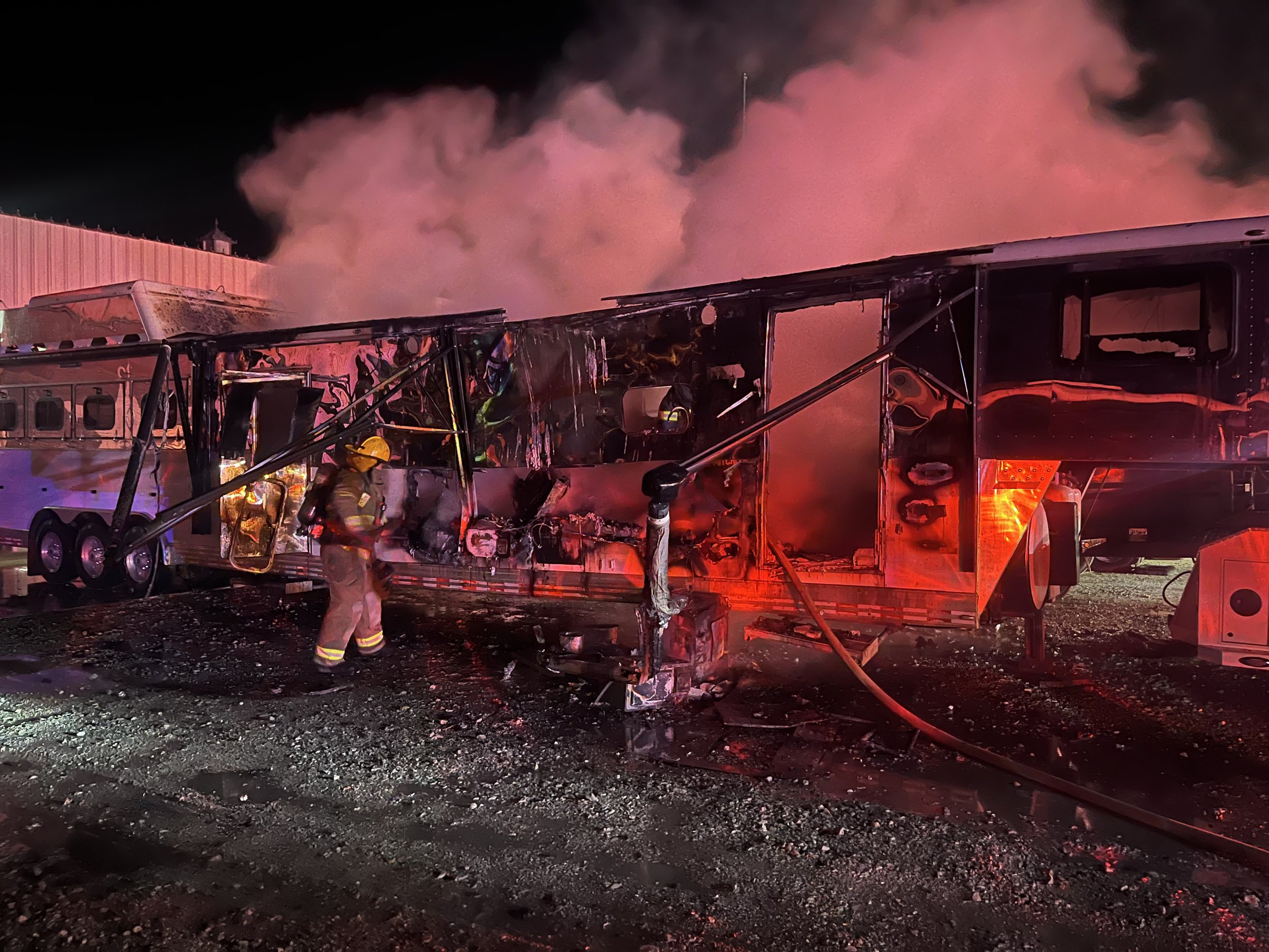 Firefighters work to extinguish a burnt-out double-decker bus at night, with smoke billowing from the scene.