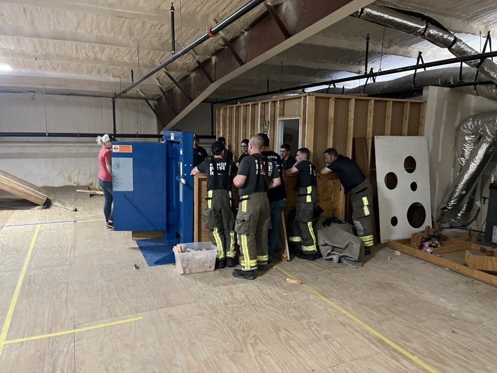 Group of firefighters assembling or inspecting a wooden structure inside a building or training facility.