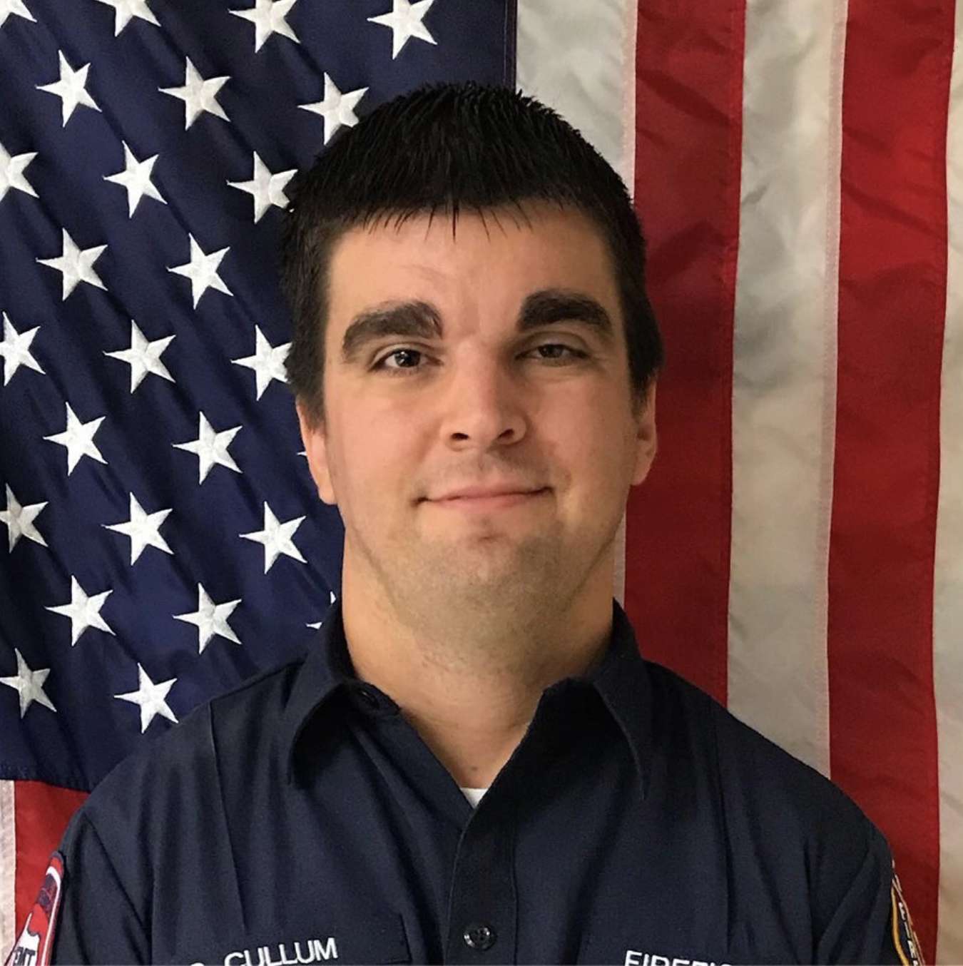A firefighter with short dark hair standing in front of an American flag, wearing a dark uniform with a badge and the name 'CULLUM' visible.