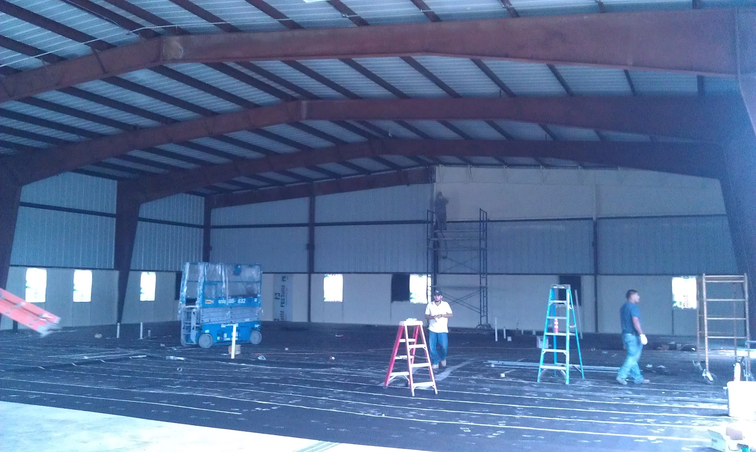 Construction workers installing radiant floor heating system inside a large warehouse or industrial building, with scaffolding, ladders, and heating pipes on the floor.