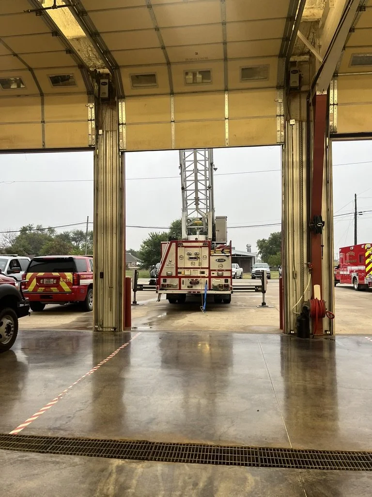 Inside a fire station garage with fire trucks and a lift, view outside shows emergency vehicles and an overcast sky.