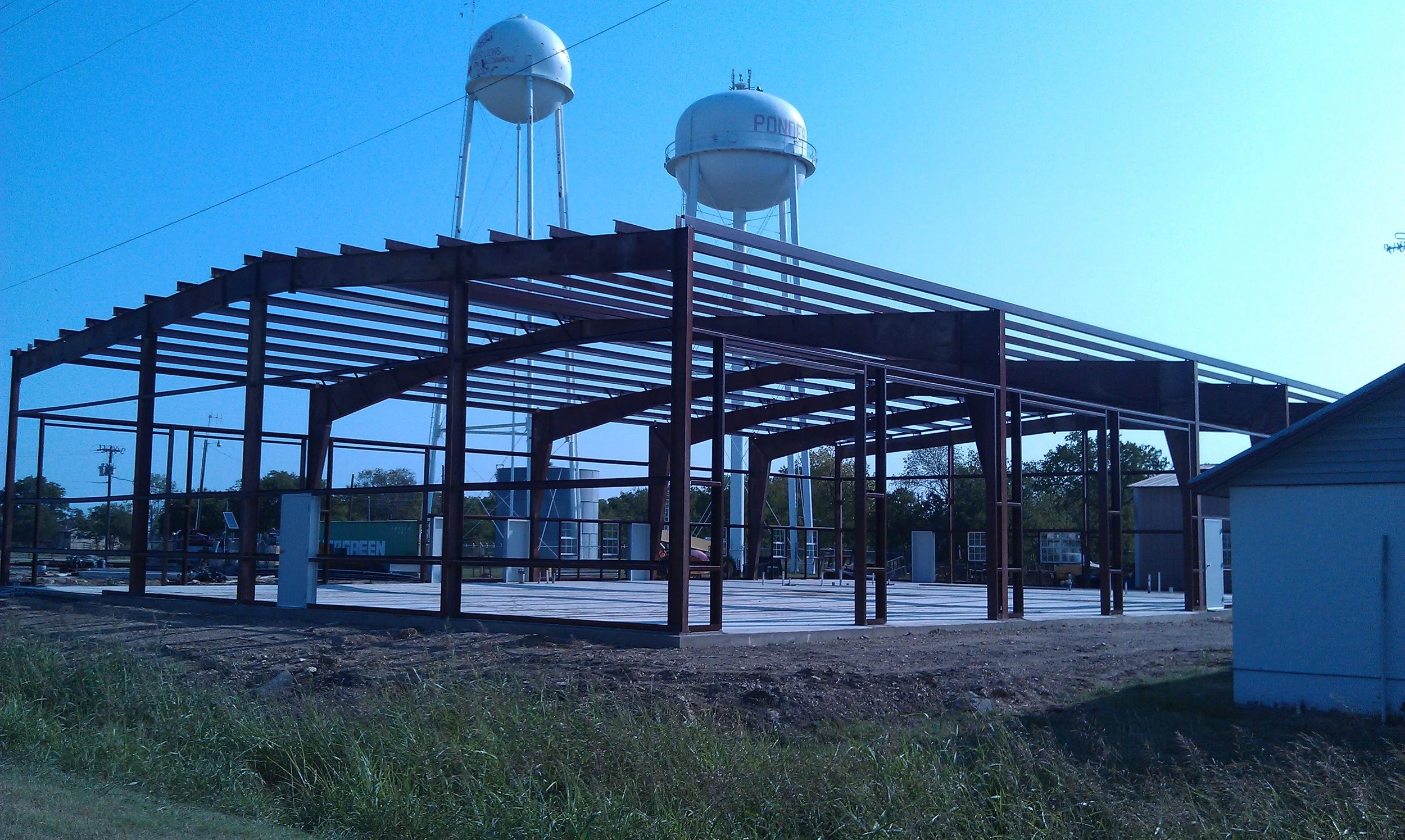 Construction site with steel framework for a building, with water towers visible in the background.