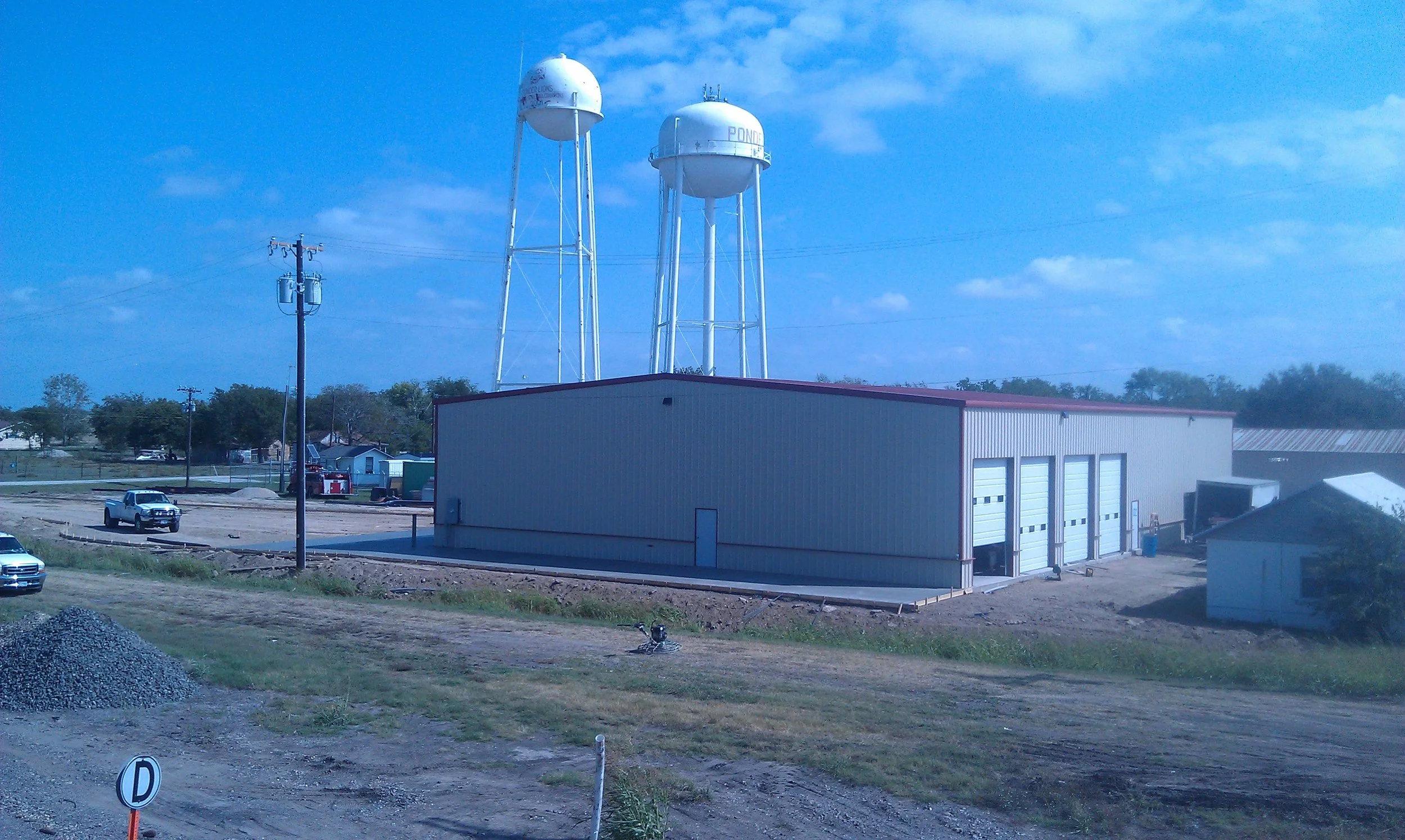 A large industrial building with multiple garage doors on a dirt lot, with two tall white water towers behind it, labeled 'POND' and 'WATER' and a clear blue sky overhead.