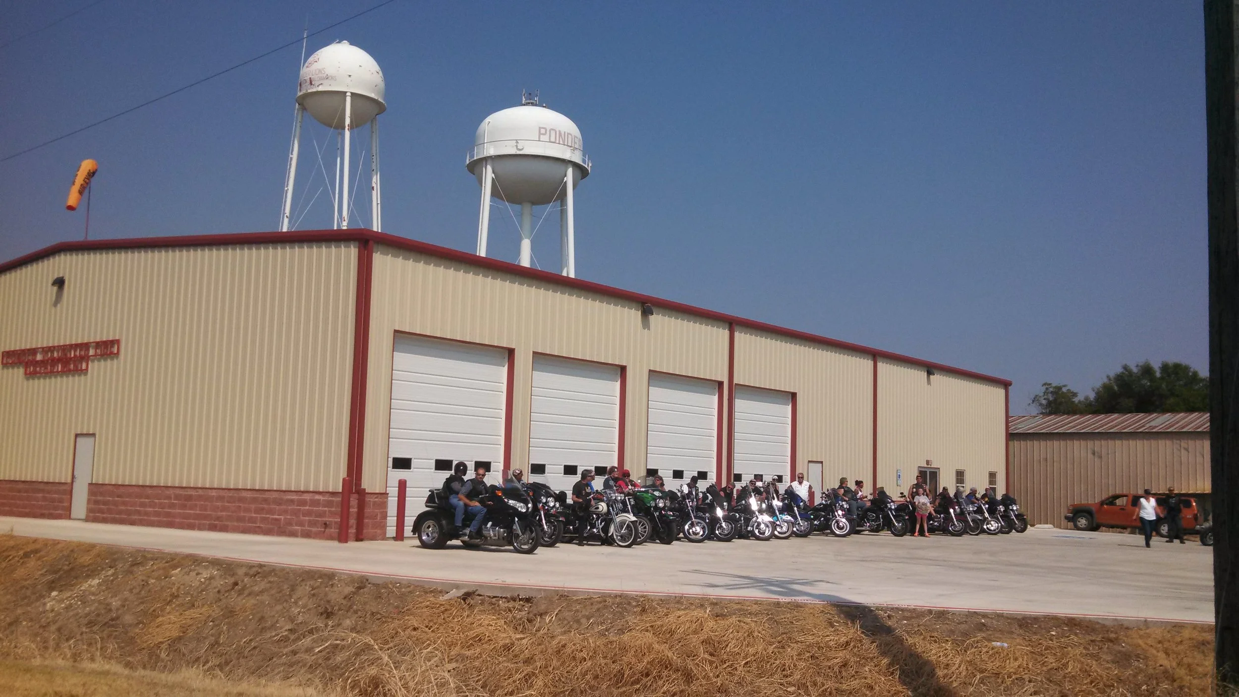 Group of motorcyclists and a person near parked motorcycles in front of a large building with garage doors, with water towers on the roof, under a clear blue sky.