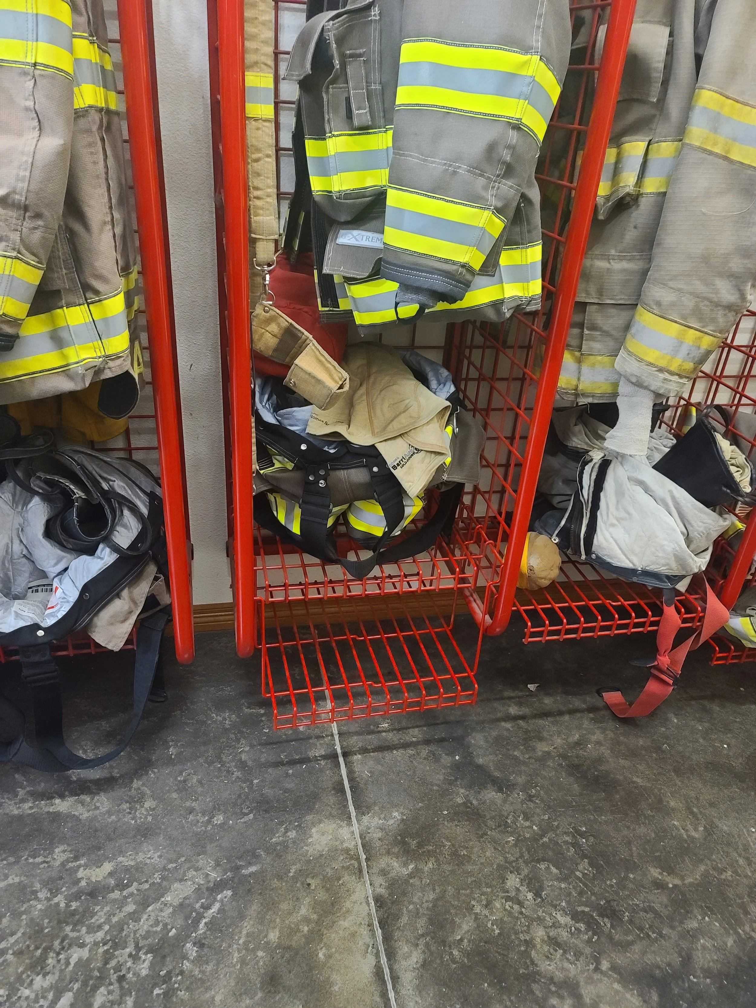 Firefighter gear including jackets, a helmet, and a bag displayed on red metal racks.