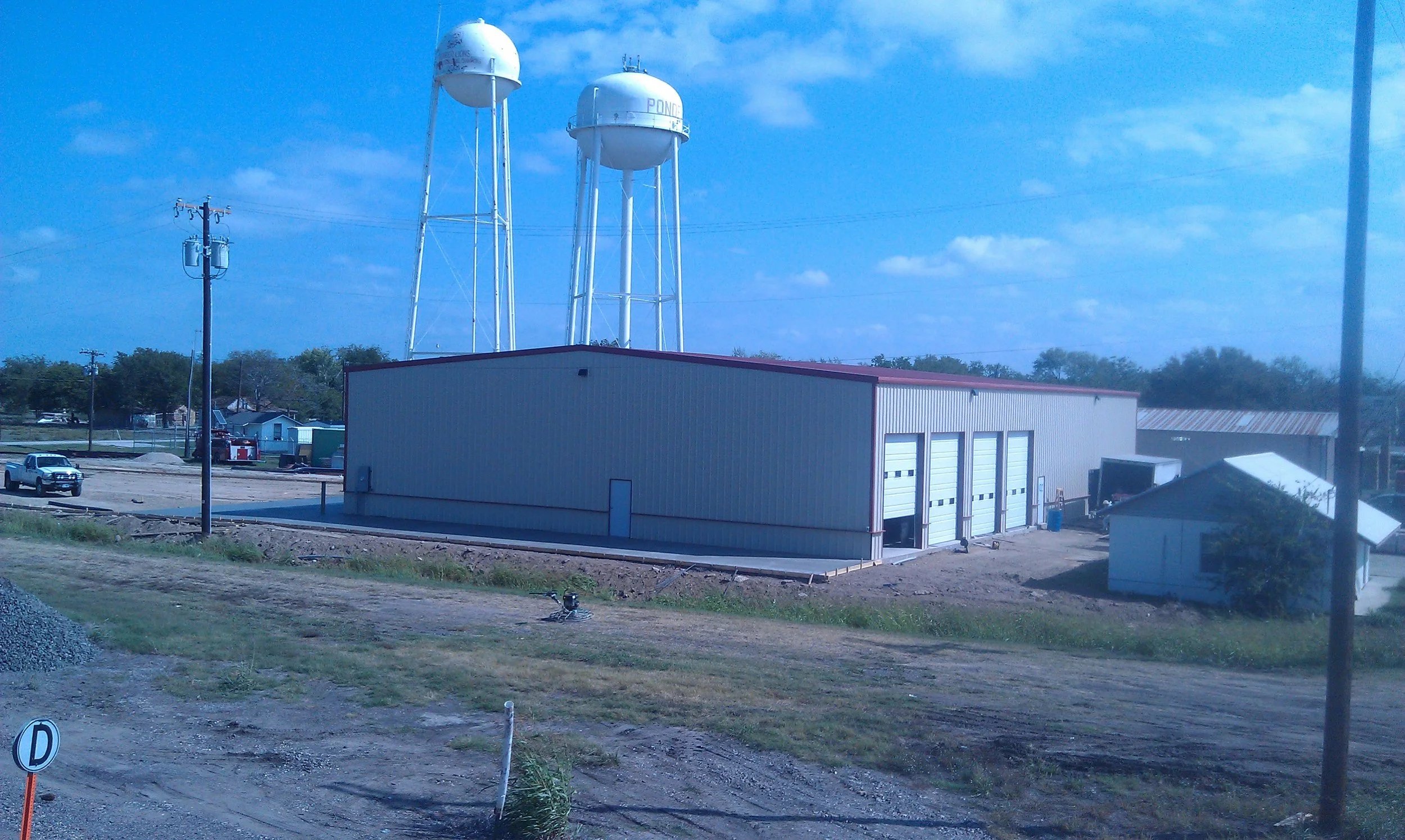 A rural scene with three water towers, a large metal building with multiple garage doors, a parked pickup truck, and a smaller building in the foreground under a partly cloudy sky.