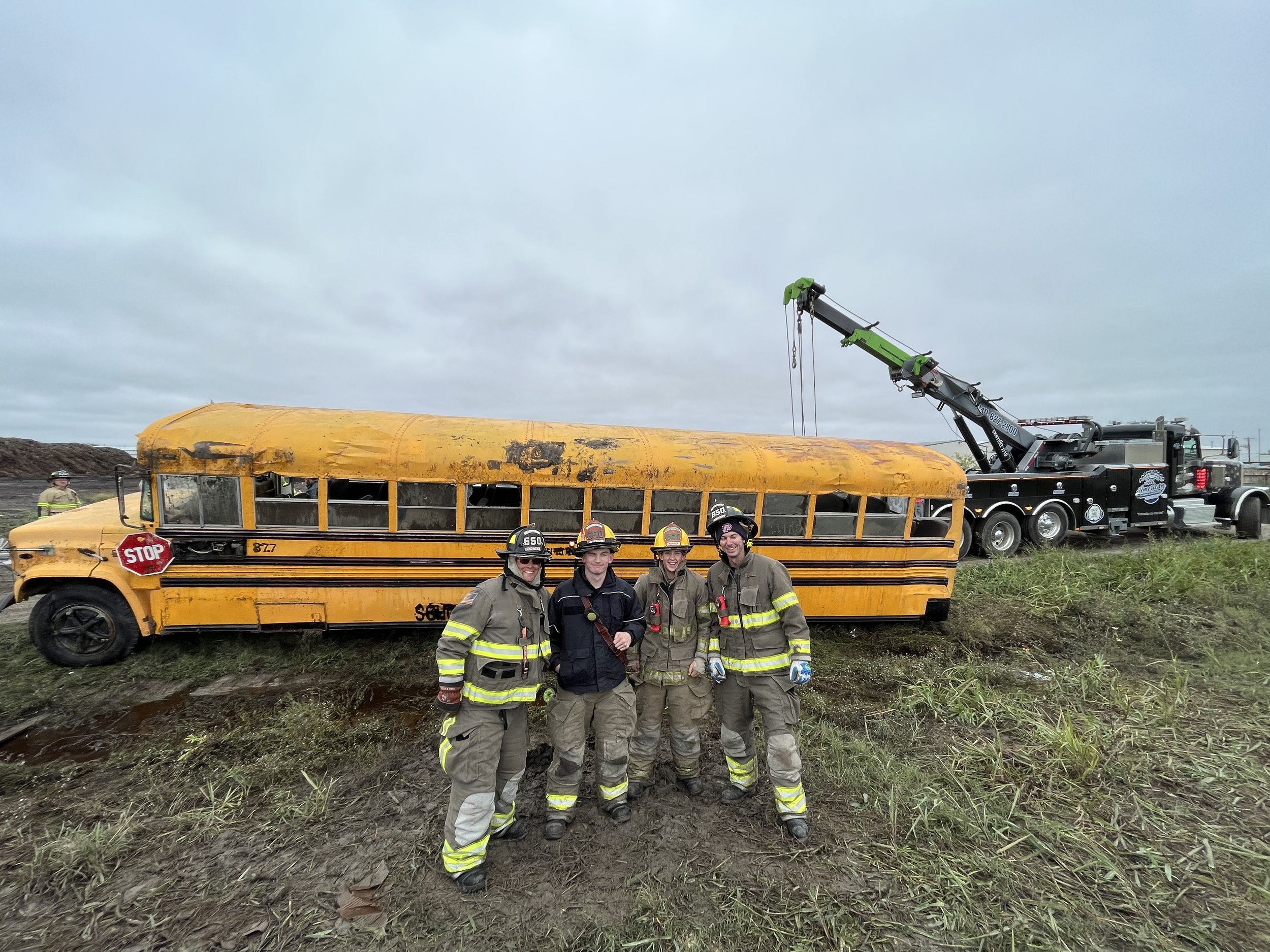 Four firefighters in uniform standing in front of a damaged yellow school bus that has been partially submerged or crashed, with a tow truck nearby in a grassy area under a cloudy sky.