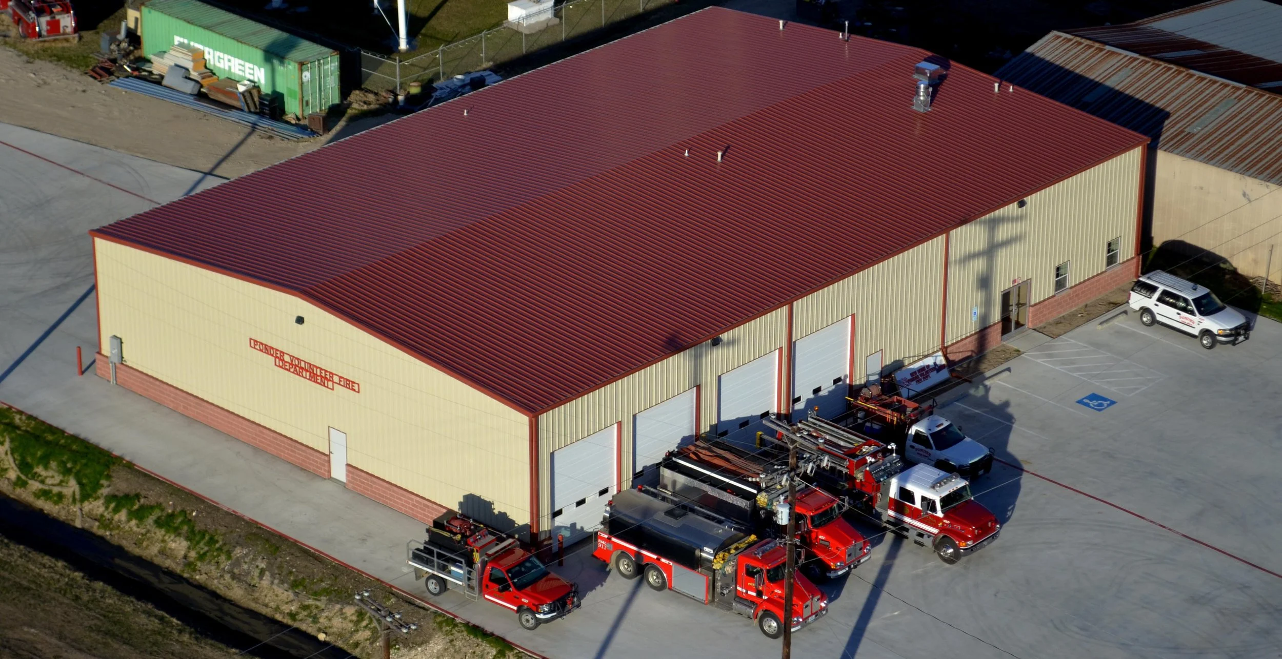 A fire department building with three fire trucks parked in front and a white pickup truck nearby. The building has a red metal roof and beige walls with a brick base, and a sign on the side indicating it is the Ponder Volunteer Fire Department.