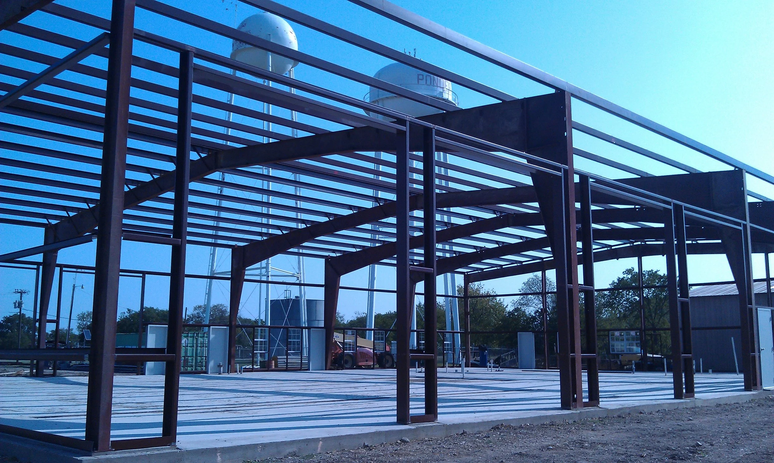 Construction site with steel framework and partially built structure, with wind turbines and utility poles in the background under clear blue sky.