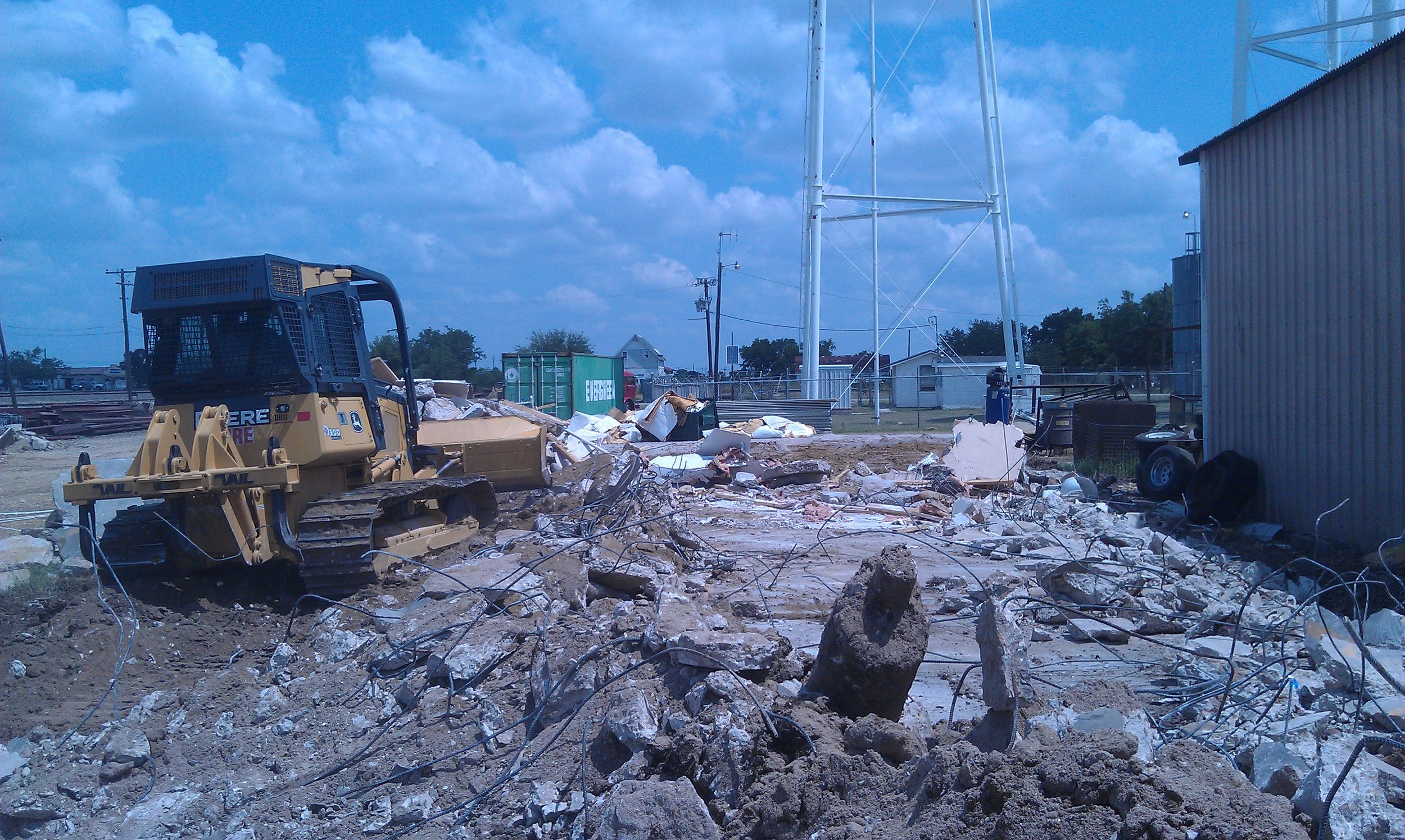 A construction site with a small bulldozer amidst rubble, debris, and bent metal wires, with a cloudy sky and power lines in the background.