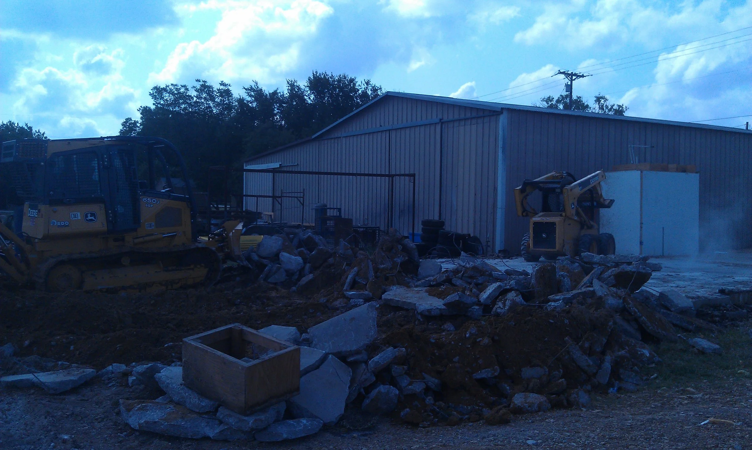 Construction site with bulldozers, rocks, and a metal building under cloudy sky.