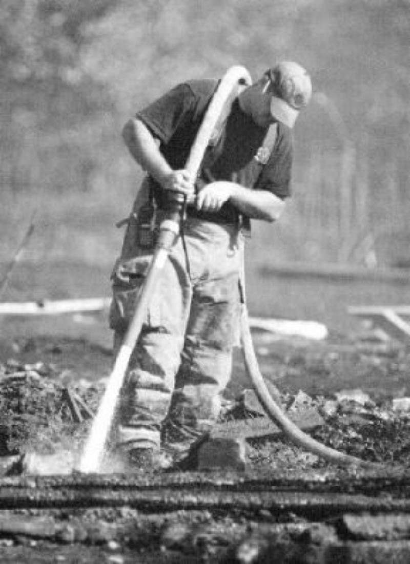A man wearing work clothes and a cap, holding a large hose, working on a construction site or road repair, with debris and signs of ongoing work around him.