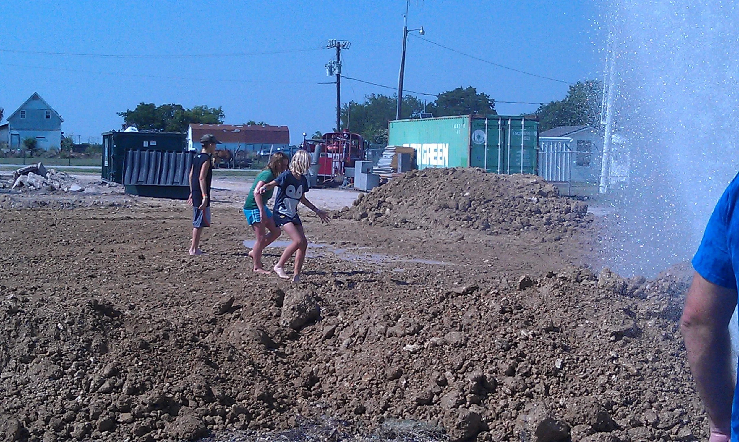 Four children playing in a muddy construction site with dirt mounds, construction equipment, and boxes in the background, some of them are barefoot.