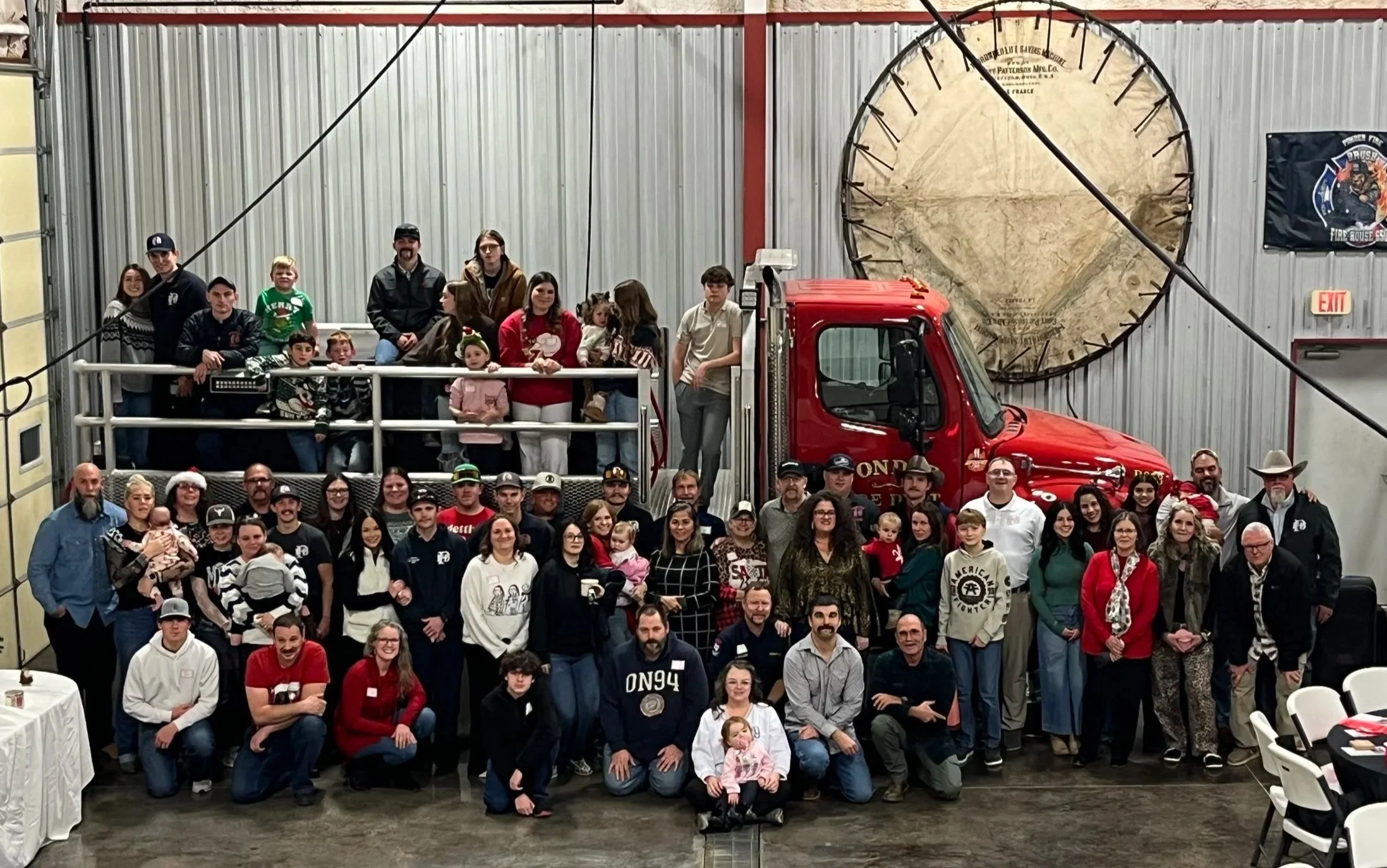 A large group of people, including children and adults, gathered around a vintage red fire truck inside a fire station for a group photo.