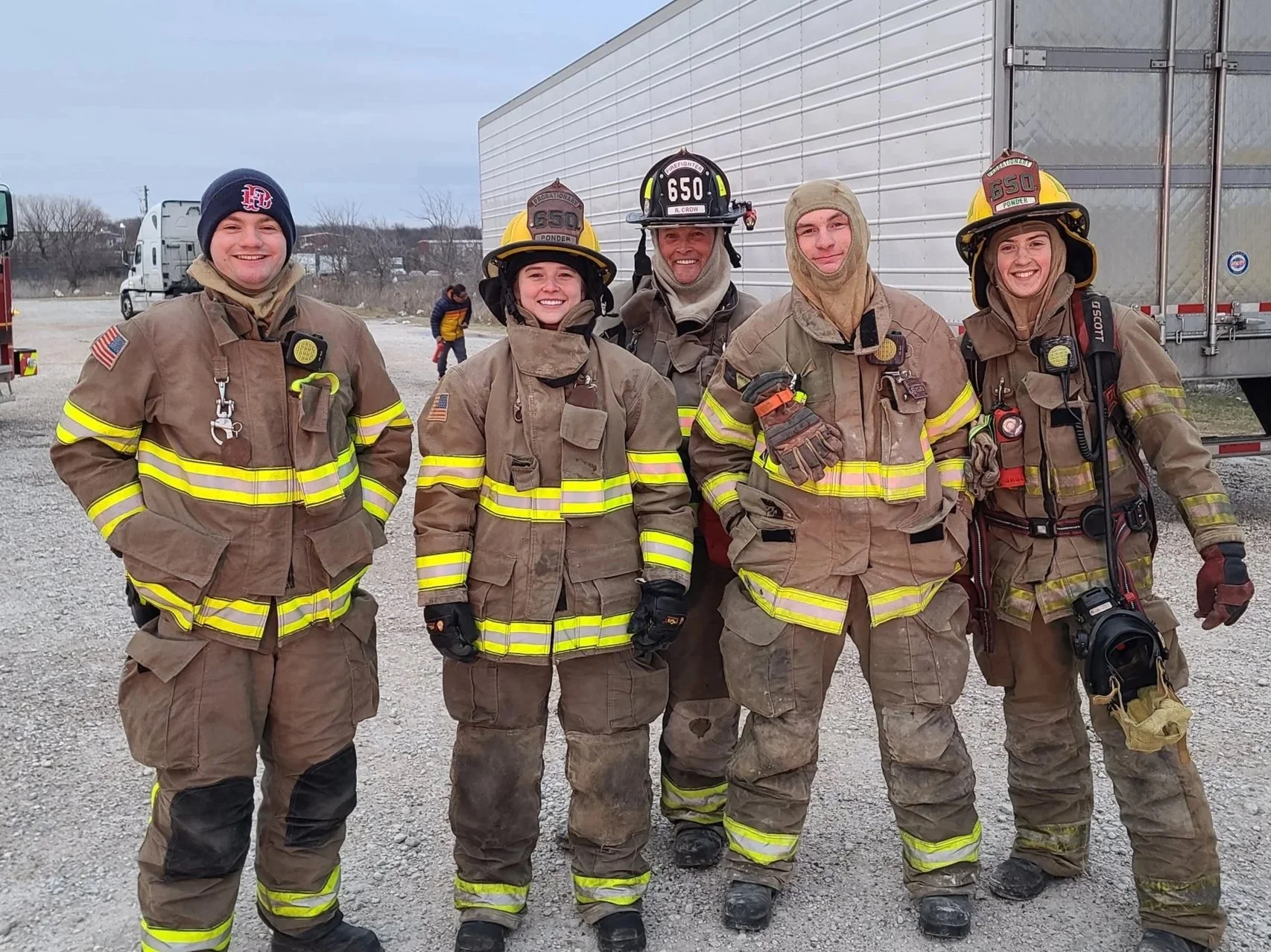 Group of five firefighters in uniform standing outdoors on a gravel area near a large truck, smiling at the camera.