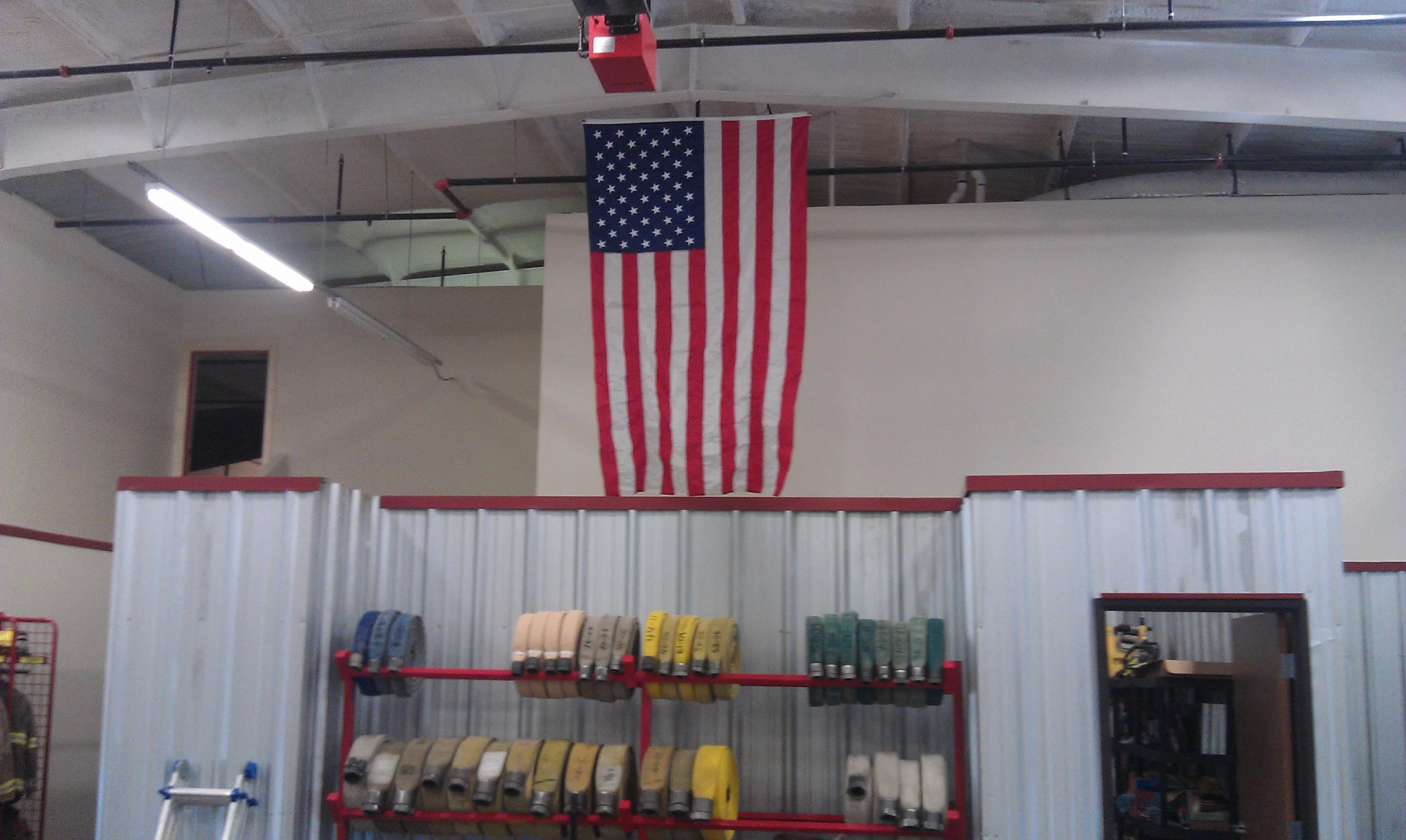Inside a fire station with a large American flag hanging from the ceiling, and organized firefighting equipment on racks in the foreground.