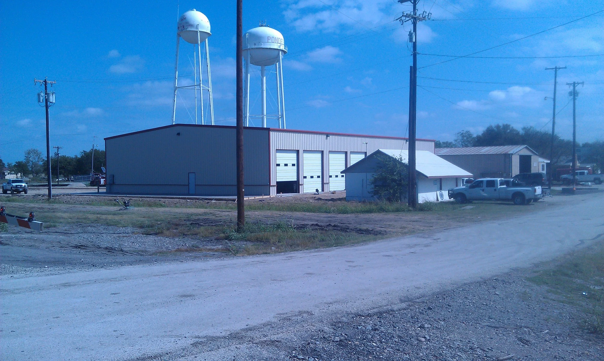 A small industrial building with three large garage doors, two water towers on metal stilts behind it, utility poles and trucks parked nearby, with a dirt road in the foreground and a blue sky with scattered clouds.