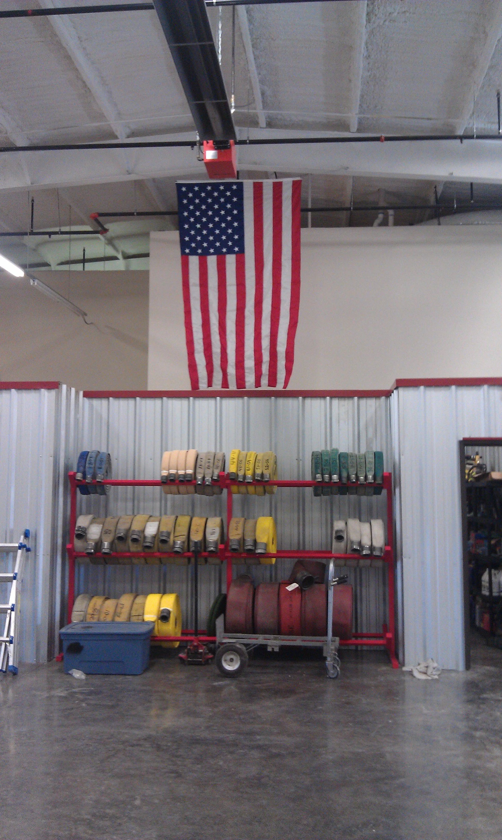 American flag hanging from the ceiling in a fire station or garage, with shelves of fire hoses and equipment below.