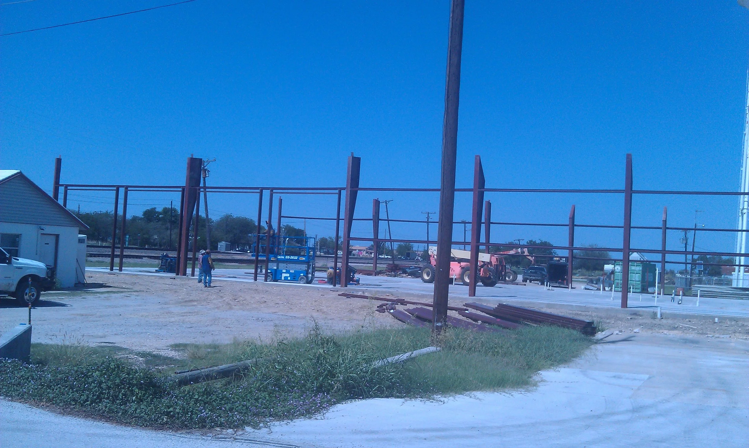 Construction site with steel framework, construction workers, machinery, parked vehicles, and a small building, under a clear blue sky.
