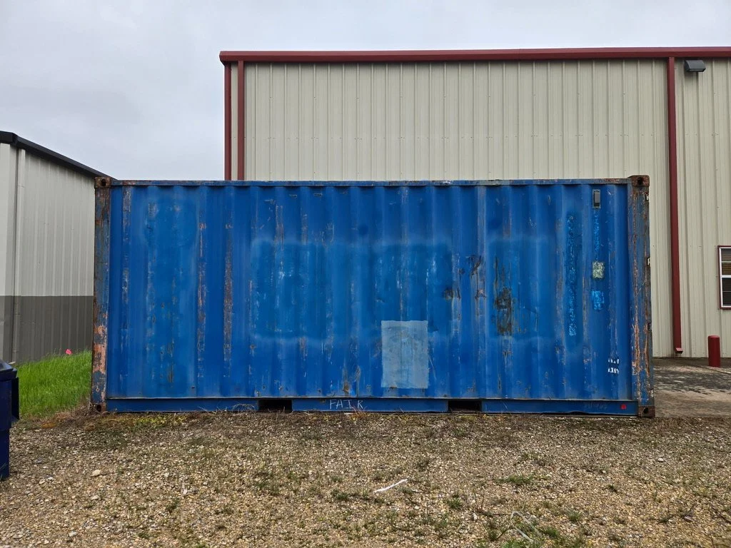 Blue weathered shipping container in front of a beige and red industrial building.