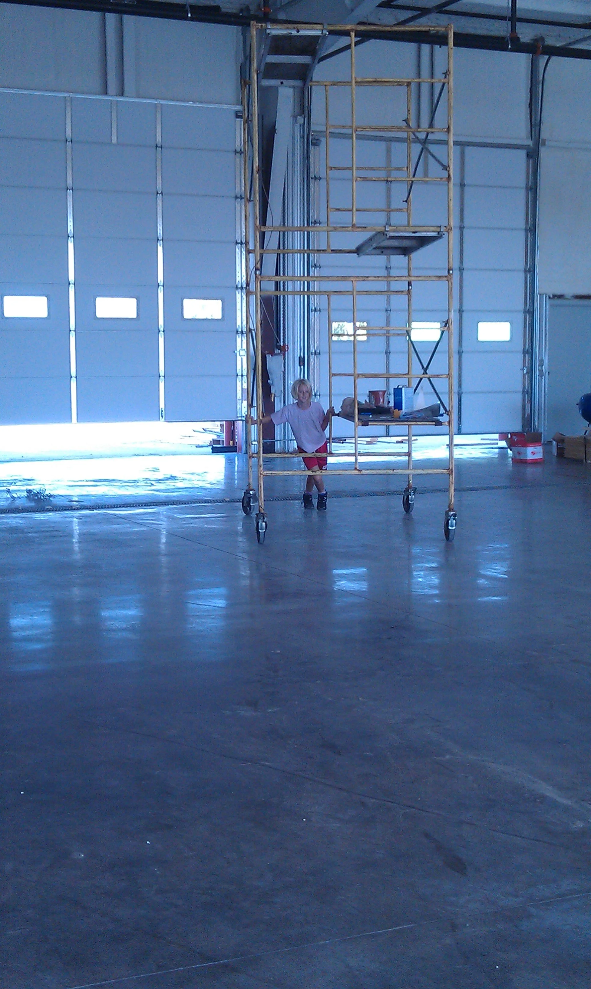 Child standing inside a wheeled scaffold in a large indoor space with open garage doors.