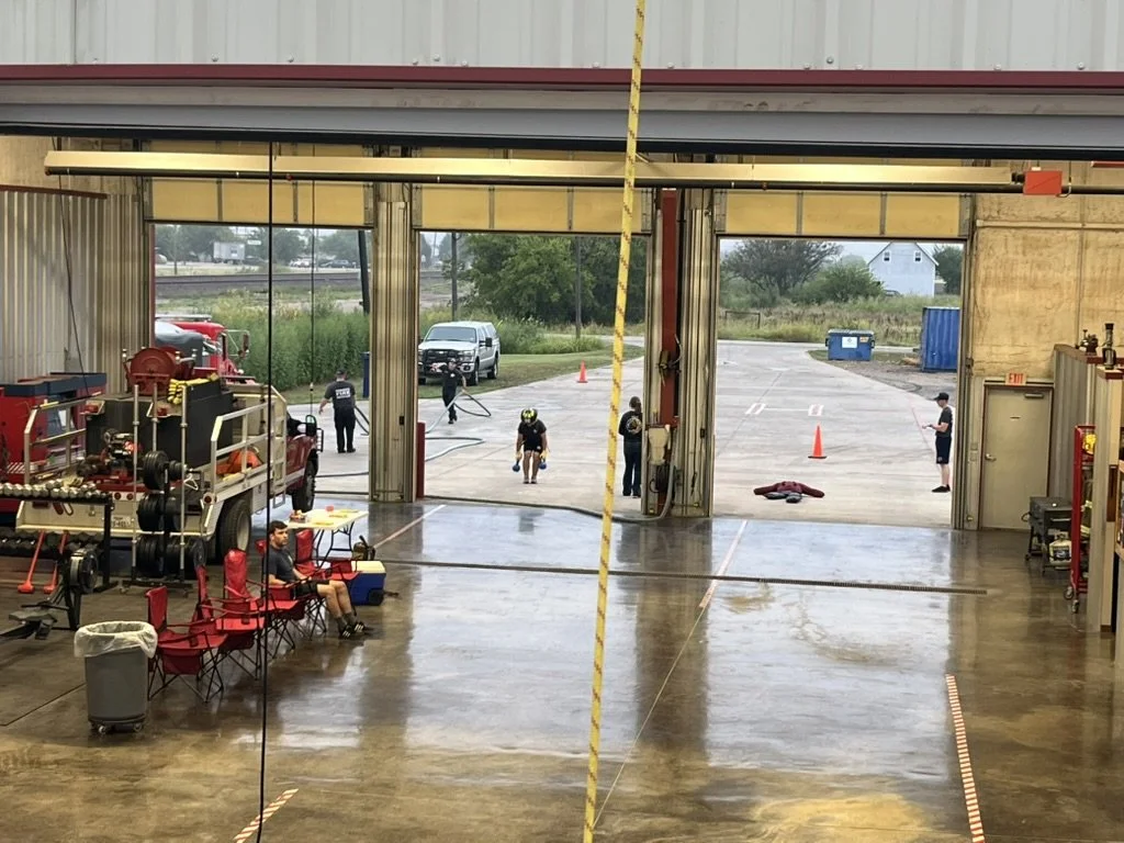 Firefighters practicing rescue drills outside a fire station garage with equipment and chairs inside.