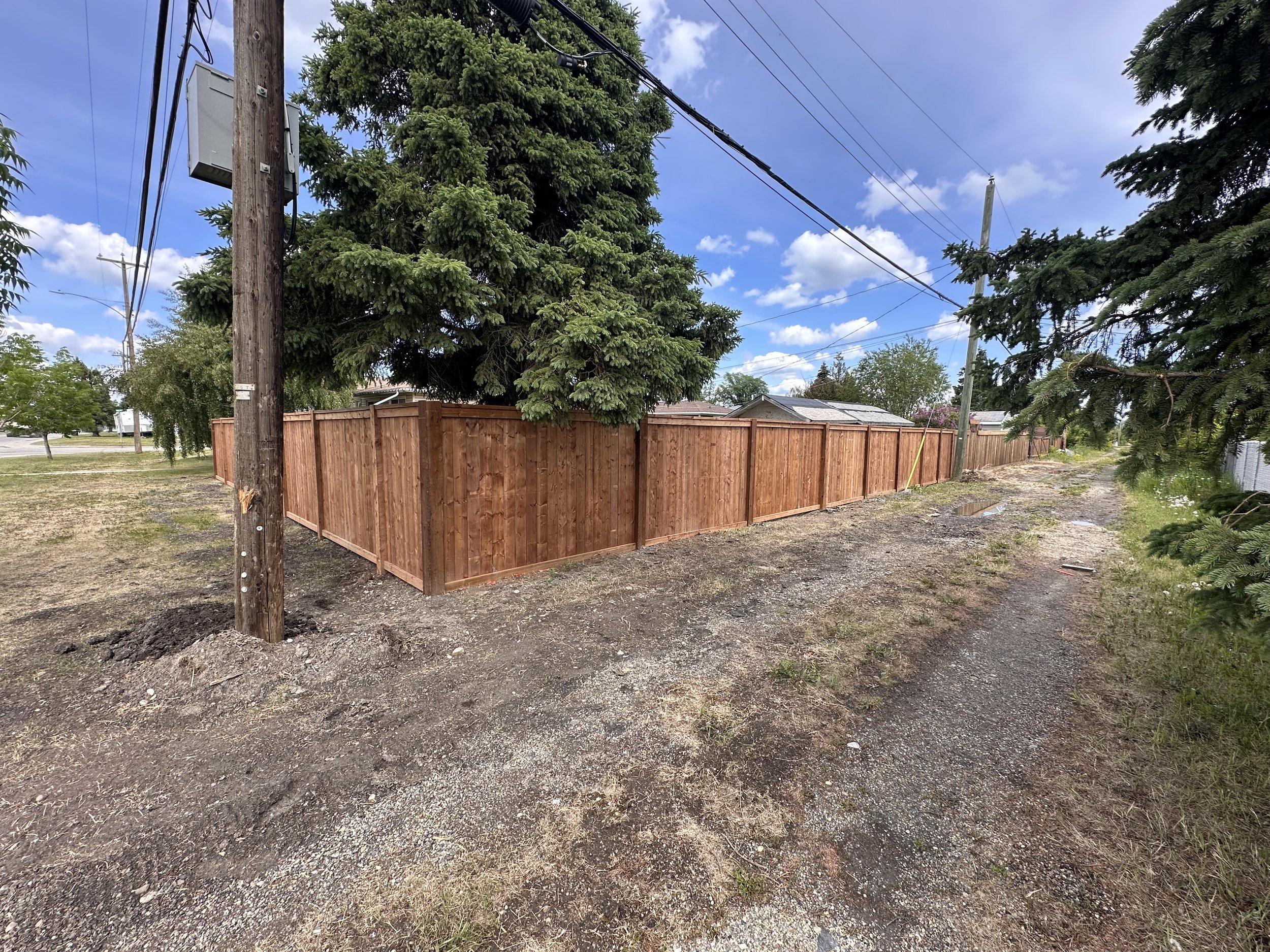 A newly built wooden fence surrounds a property, with large evergreen trees behind it. Gravel and dirt path runs alongside the fence, under a partly cloudy sky.