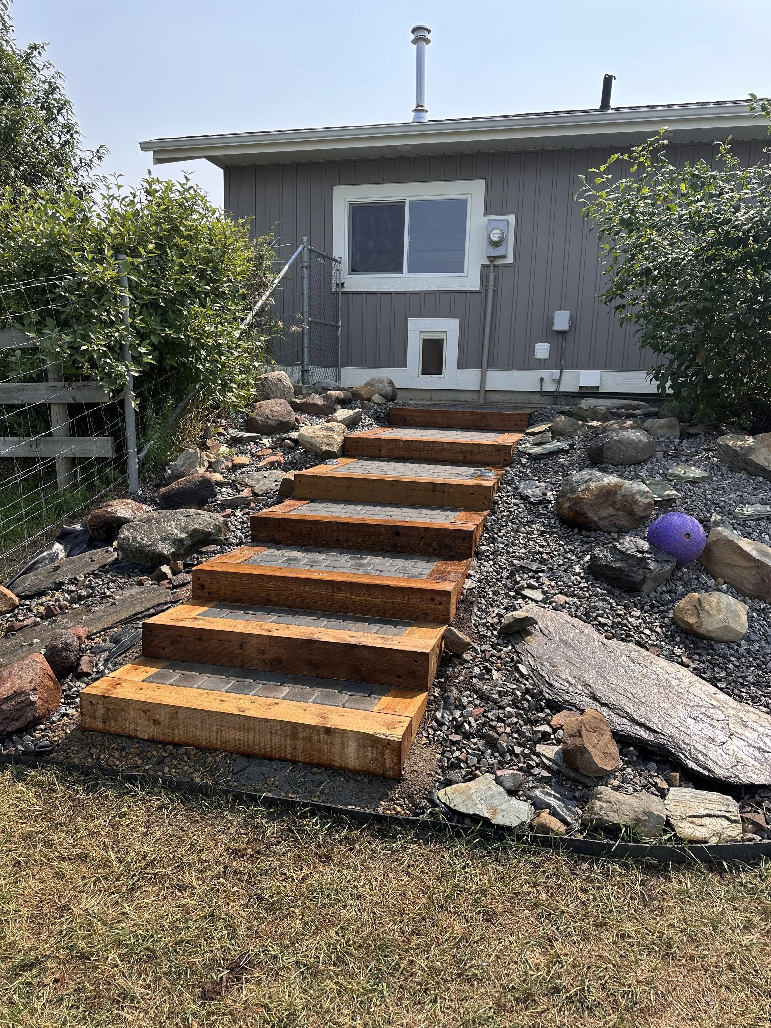 Newly built wooden stairs leading up a rocky, landscaped yard toward a house with gray siding, a window, and a small door.
