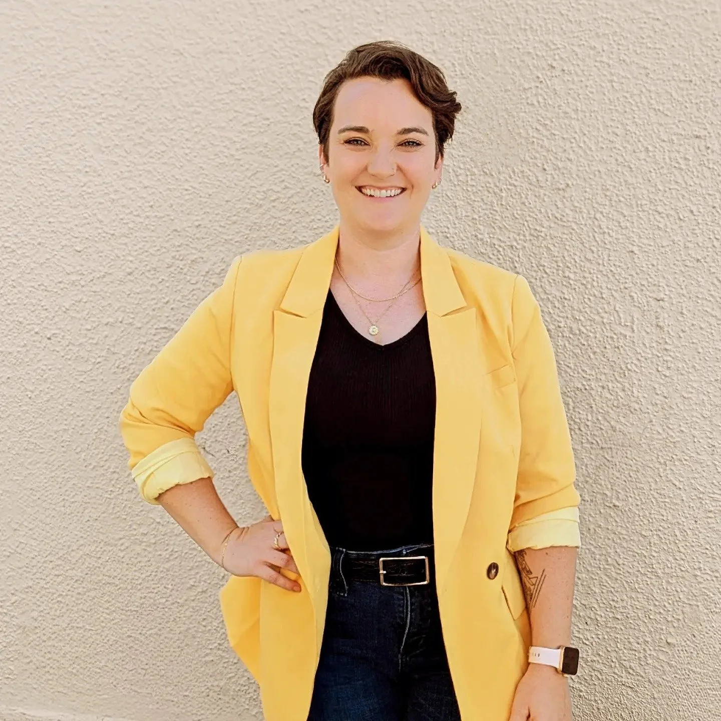 Smiling woman in a yellow blazer and black outfit standing against a textured wall